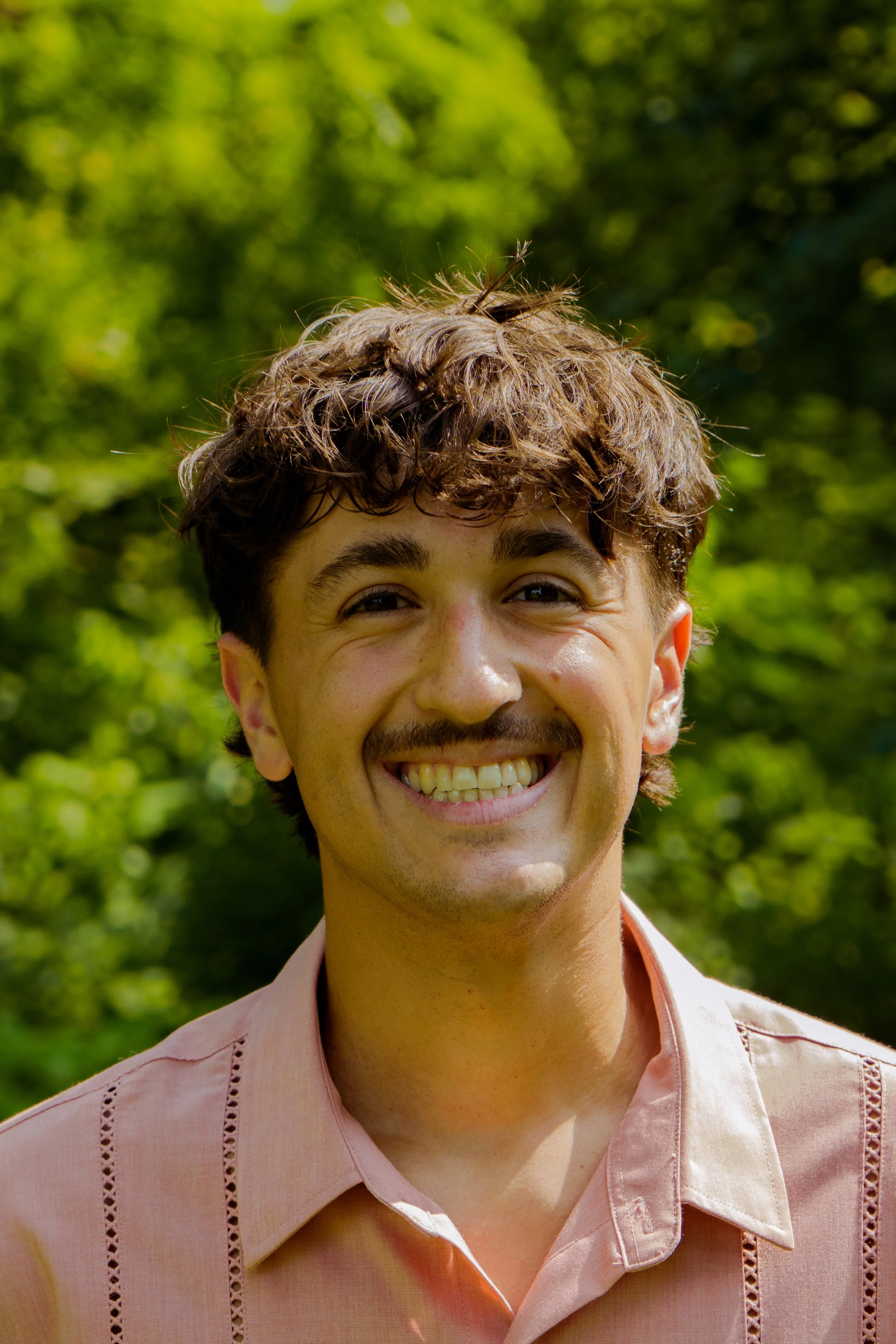 A young man with curly brown hair and a mustache, smiling, outdoors with green trees in the background, wearing a light pink button-up shirt.
