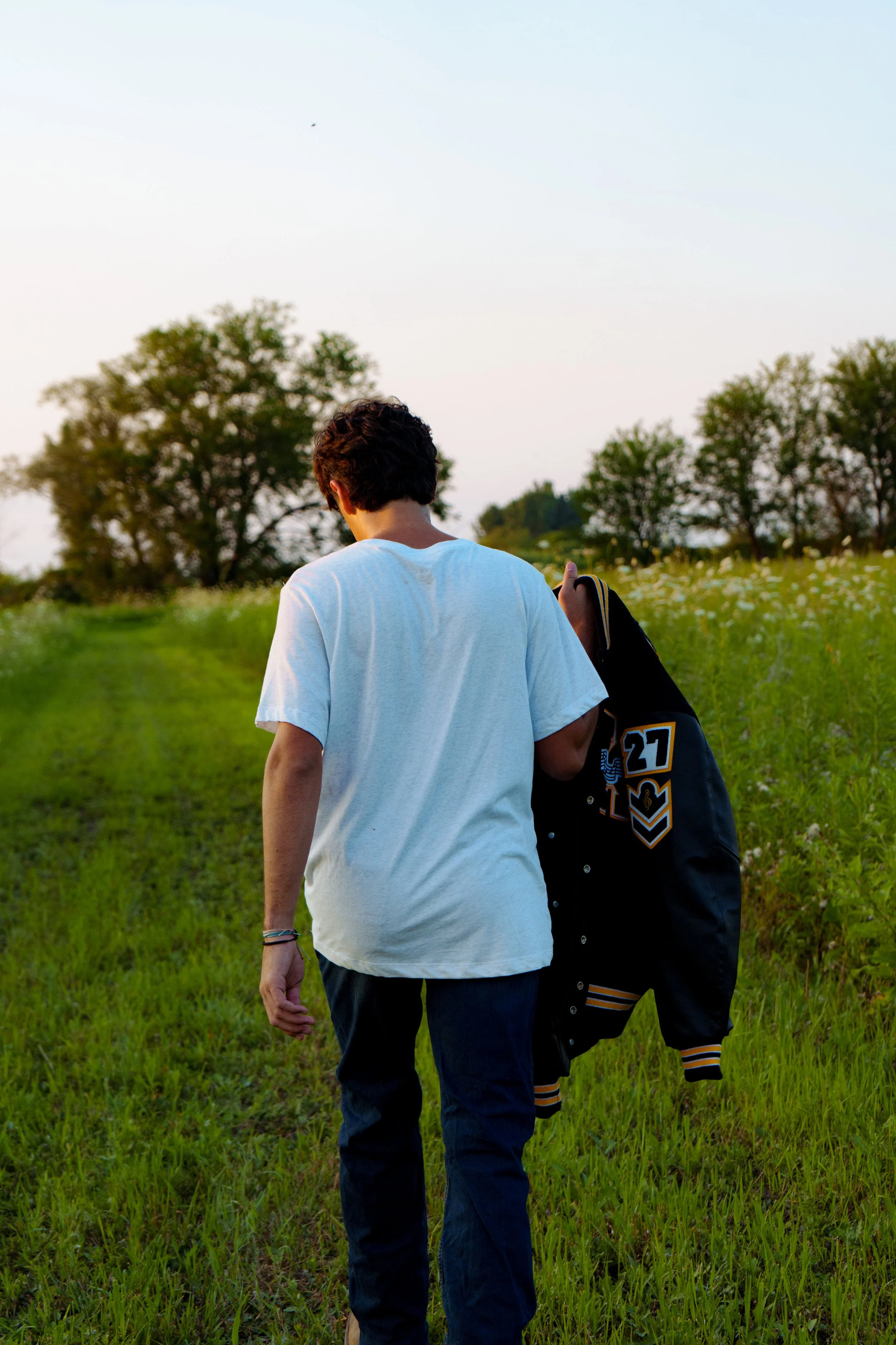 A person with dark curly hair, wearing a white T-shirt and jeans, walking through a grassy field at sunset, carrying a black and yellow sports bag with the number 27 on it.