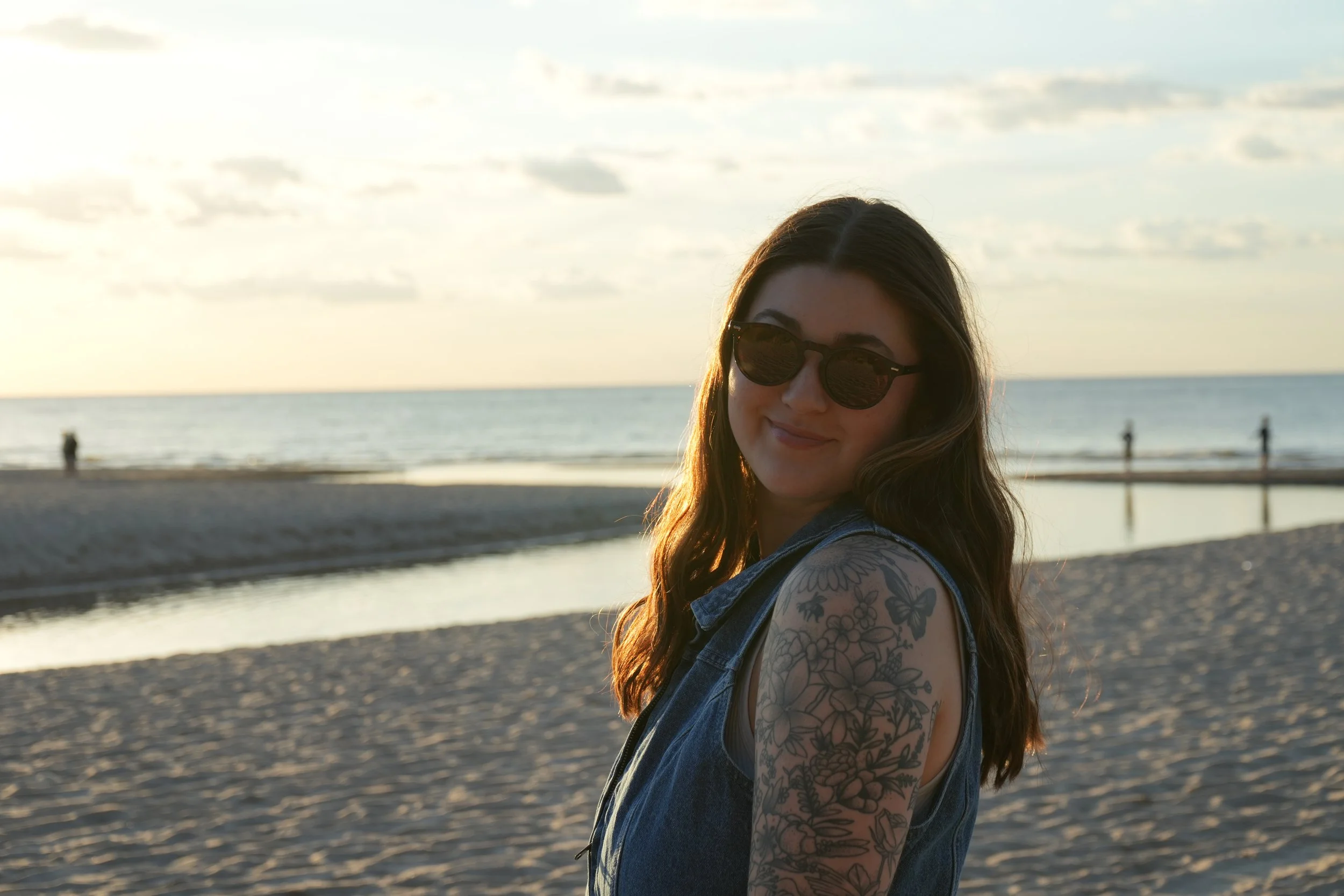 A woman with long wavy hair, wearing sunglasses and a sleeveless denim jacket, smiling at the camera, standing on a beach during sunset with an ocean and a few people in the background.