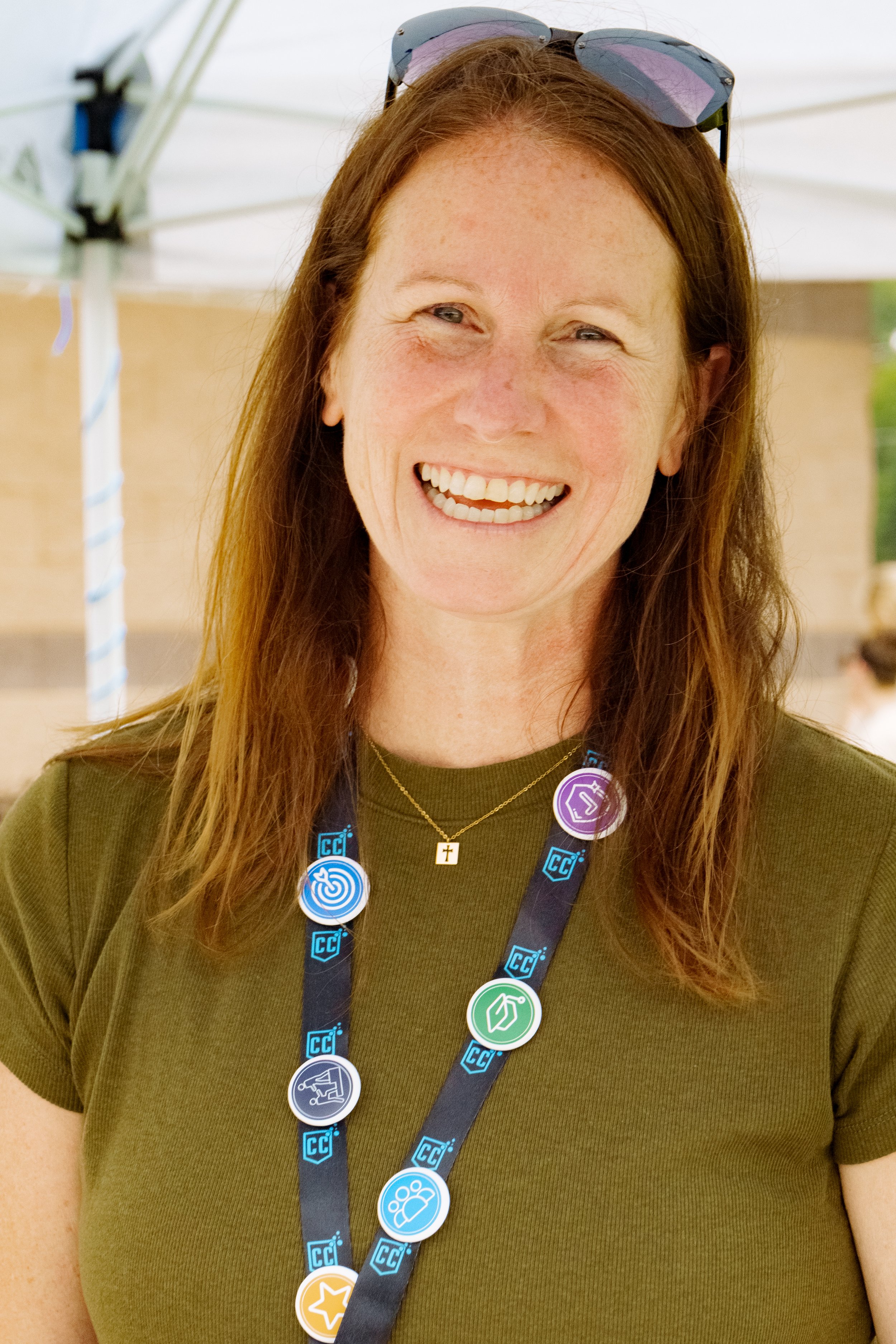 A woman with brown hair wearing sunglasses on her head, smiling, and wearing different pins on a lanyard around her neck, standing in front of a white tent outdoors.