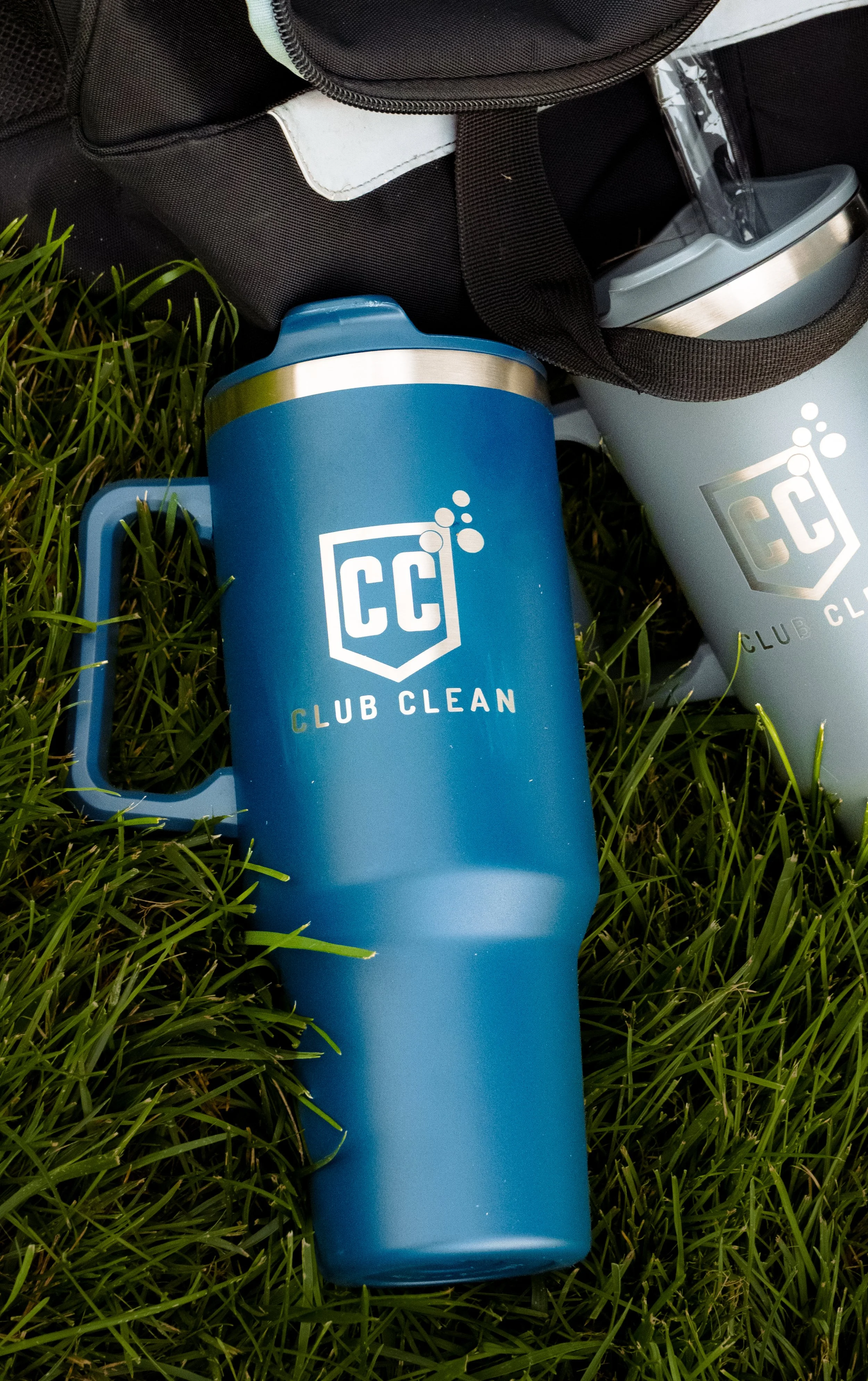 Two insulated tumblers with a 'Club Clean' logo on a grassy surface, one blue and one gray, partially inside a black carrying case.