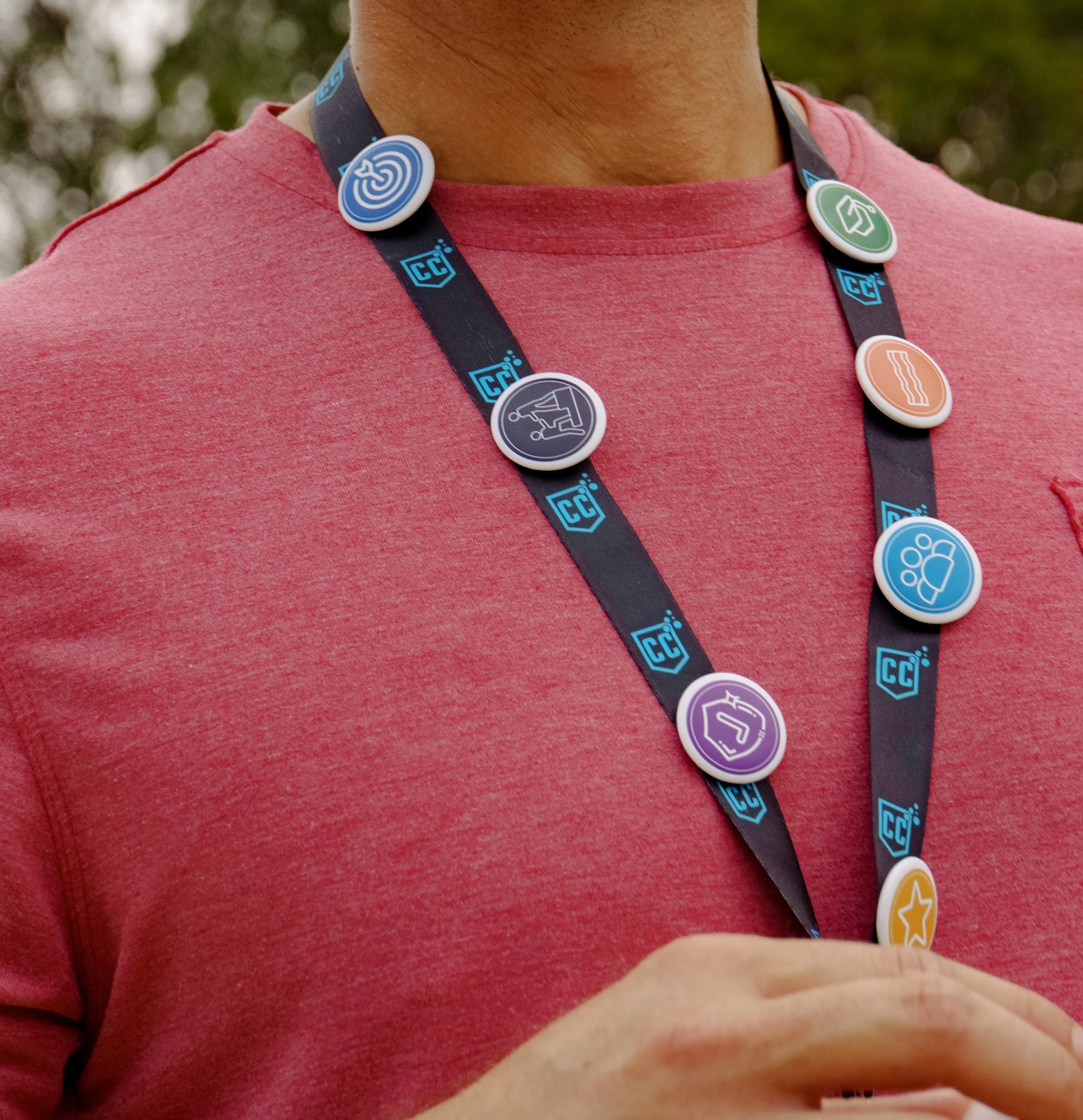 Close-up of a person wearing a red shirt and a lanyard with various colorful circular icons, including a target, house, showerhead, and shower, hanging around their neck.