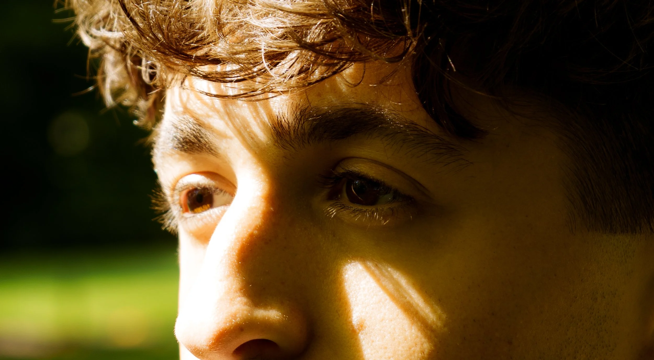 Close-up of a young person's face with short, wavy brown hair, brown eyes, and smooth skin, partially illuminated by sunlight against a green background.