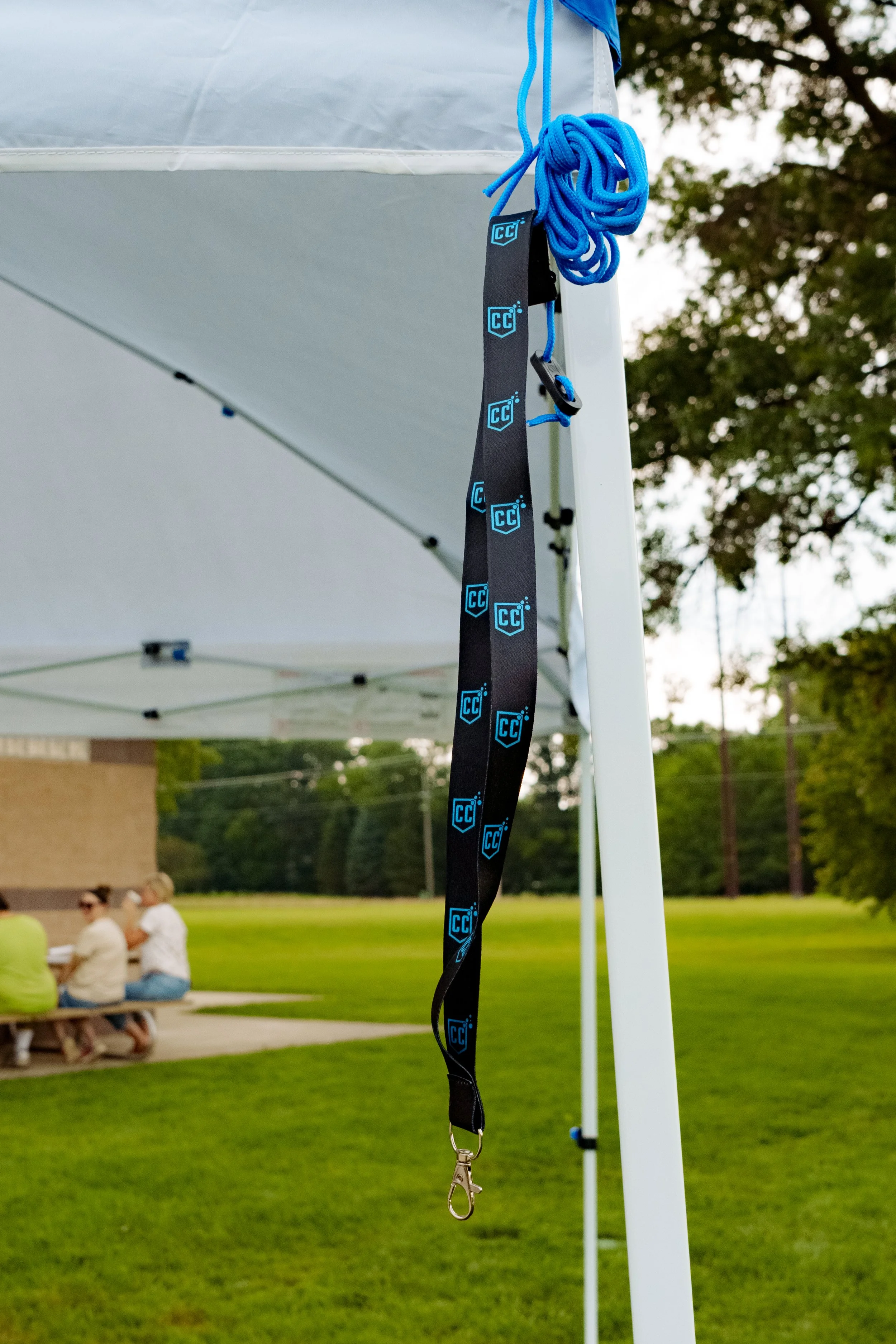 A black lanyard with blue logo hanging from the corner of a white canopy tent at a park or outdoor event.