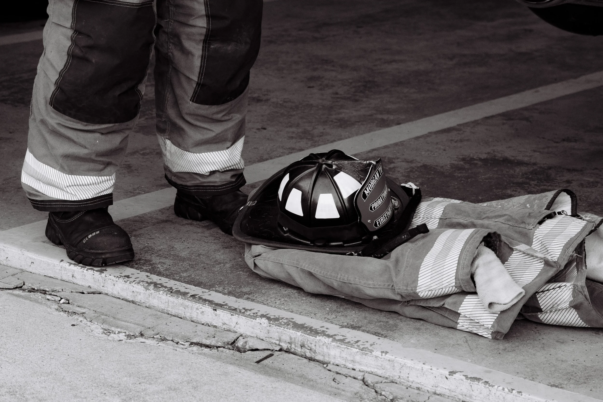 A firefighter is standing next to a person lying on the ground, with a firefighter helmet and gear placed on the person's chest. The firefighter is wearing black boots and protective pants, and is on a concrete surface, possibly a parking lot.