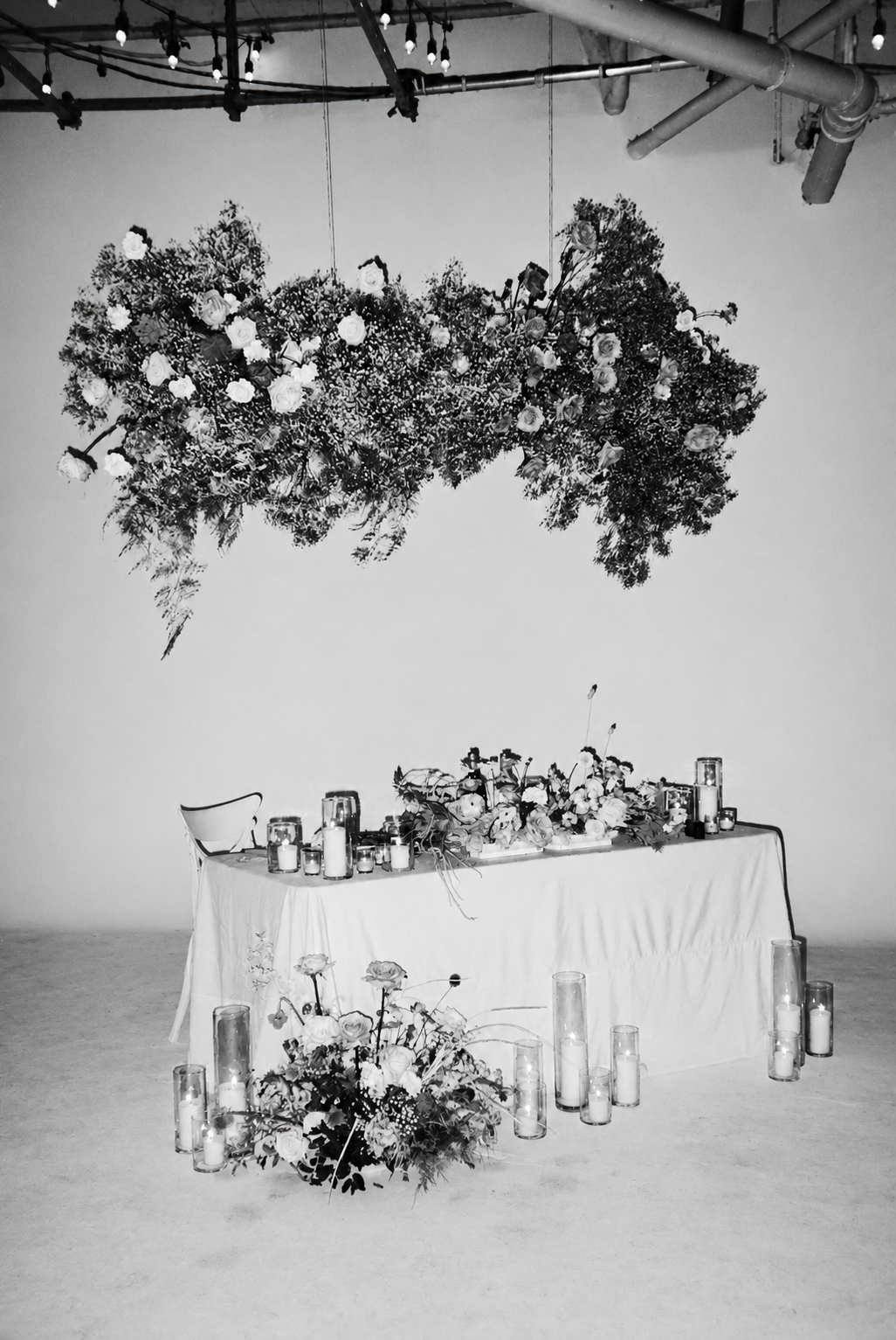 A decorated table with floral arrangements and candles, and a large floral installation hanging from the ceiling in a minimalist setting.
