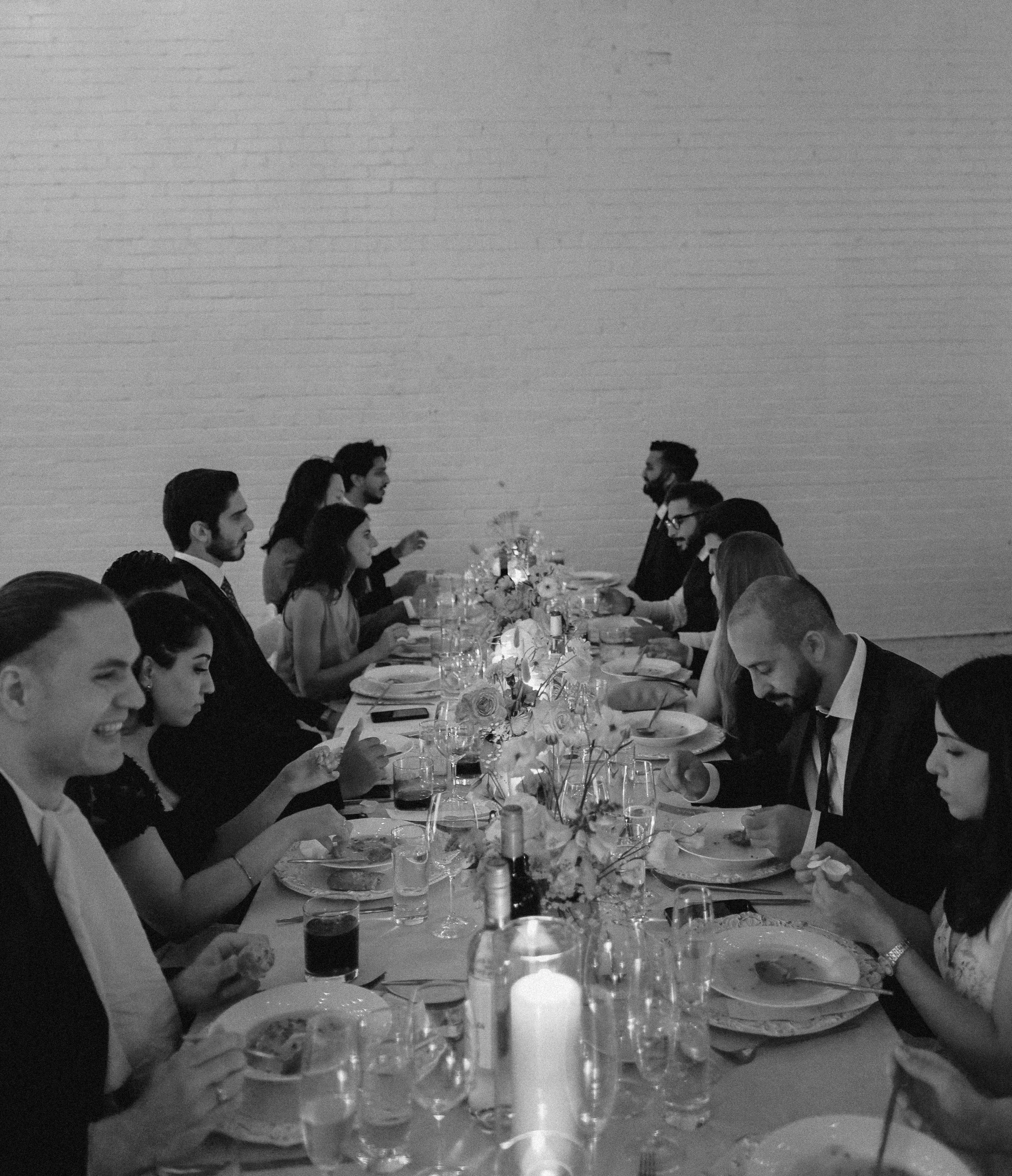 People sitting at a long dining table, dressed formally, having a meal in a decorated setting with flowers and candles.