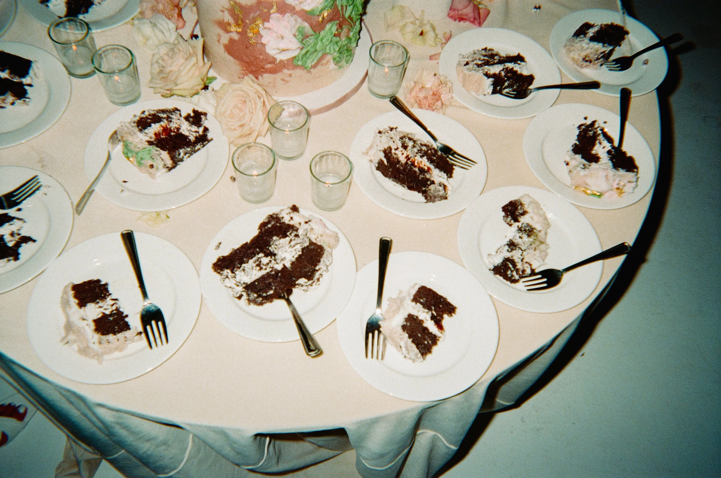 Several slices of chocolate cake with cream on white plates, set on a round table with a floral centerpiece and empty glasses.