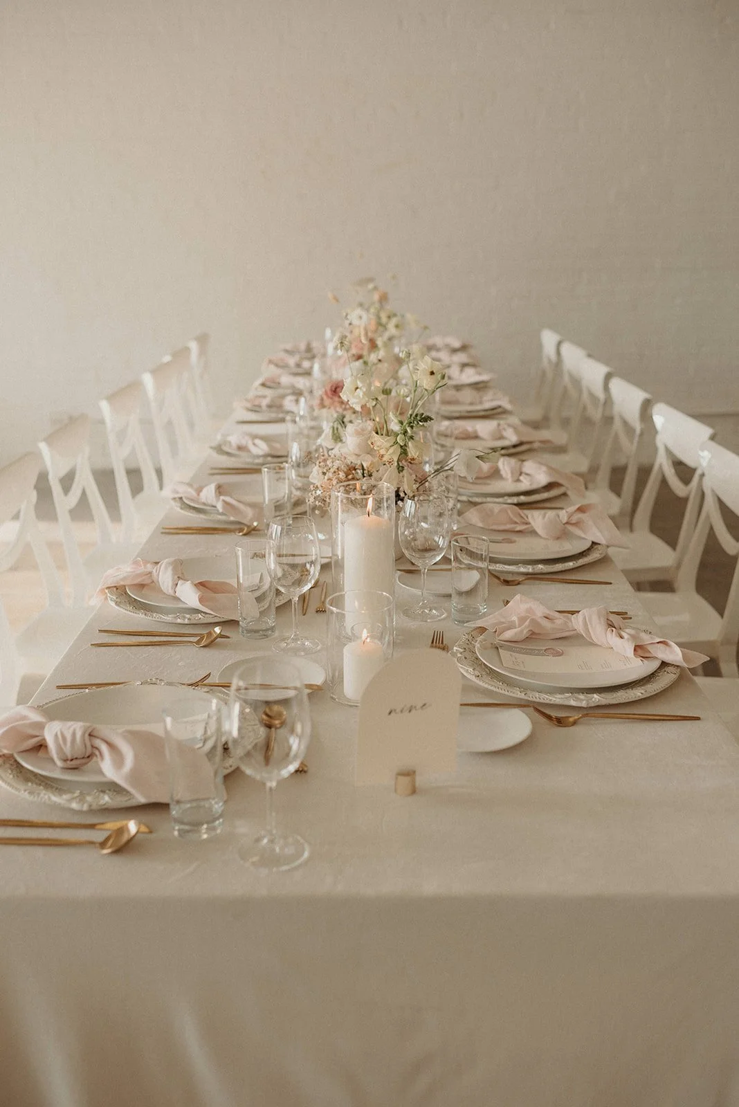 Elegant tablesetting for a formal event with pink flowers, candles, gold utensils, and pastel napkins on a long white table.