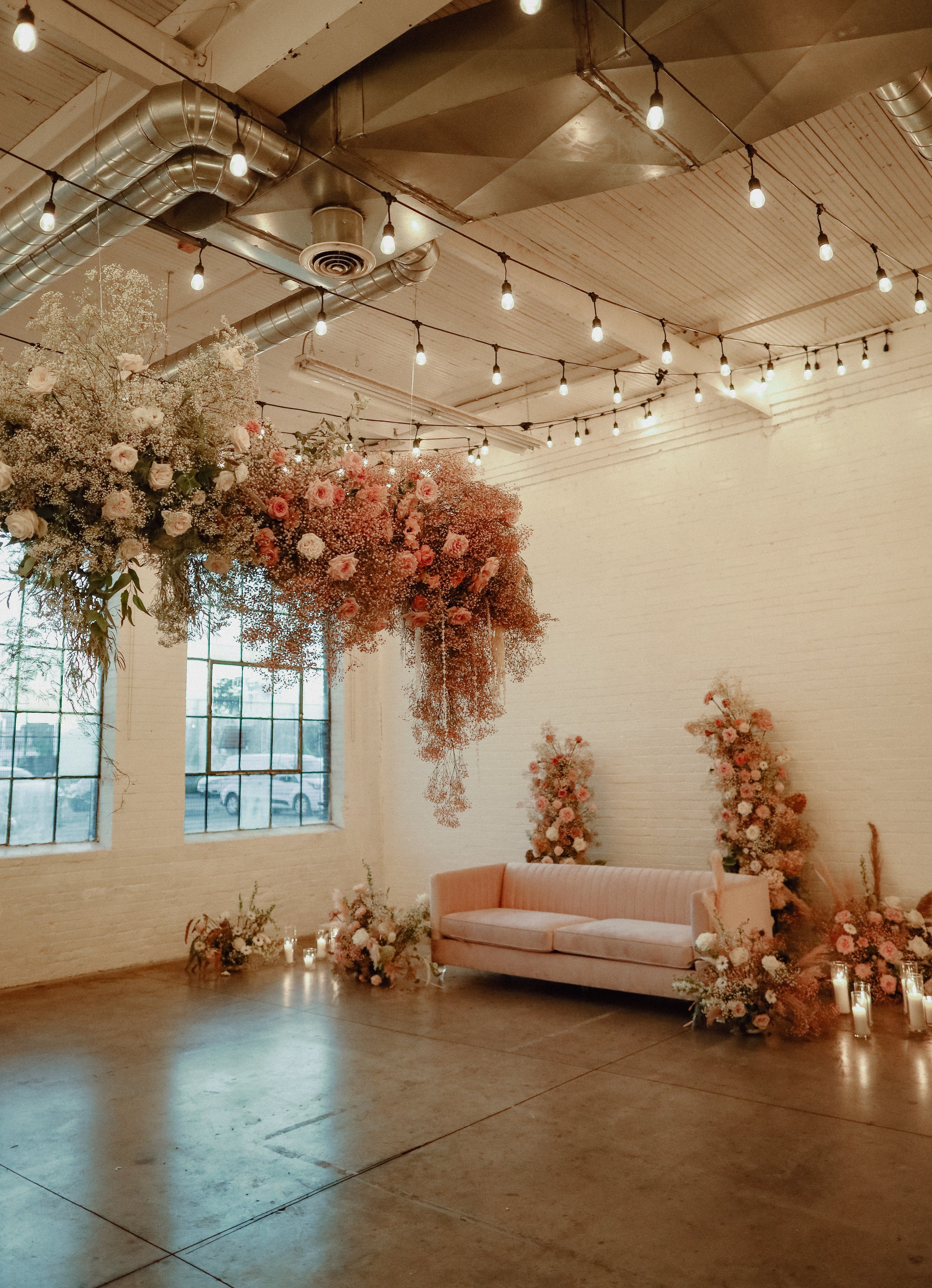 Indoor wedding or event space decorated with a large hanging floral arrangement of pink and white roses and baby's breath. Soft pink sofa with lit candles, and tall floral displays with pink roses in the background. Exposed ceiling with string lights and industrial vents.