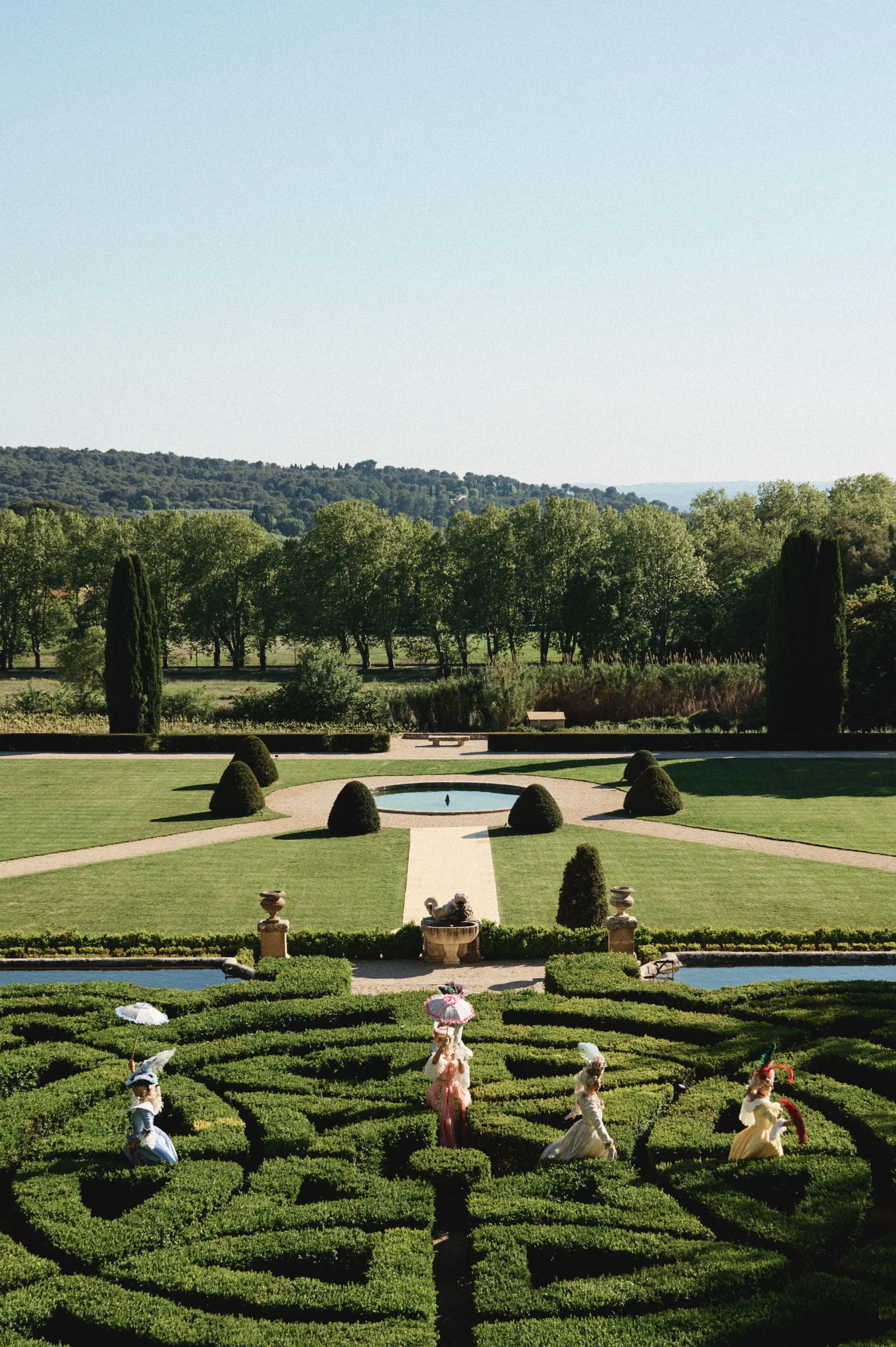 A manicured garden with a symmetrical maze of bushes, small statues, and a fountain, with lush green trees and hilly landscape in the background, and a clear sky above.