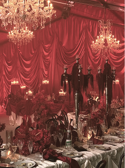 An elegant banquet hall decorated with red drapes, multiple chandeliers, and ornate table settings with candles and floral centerpieces.