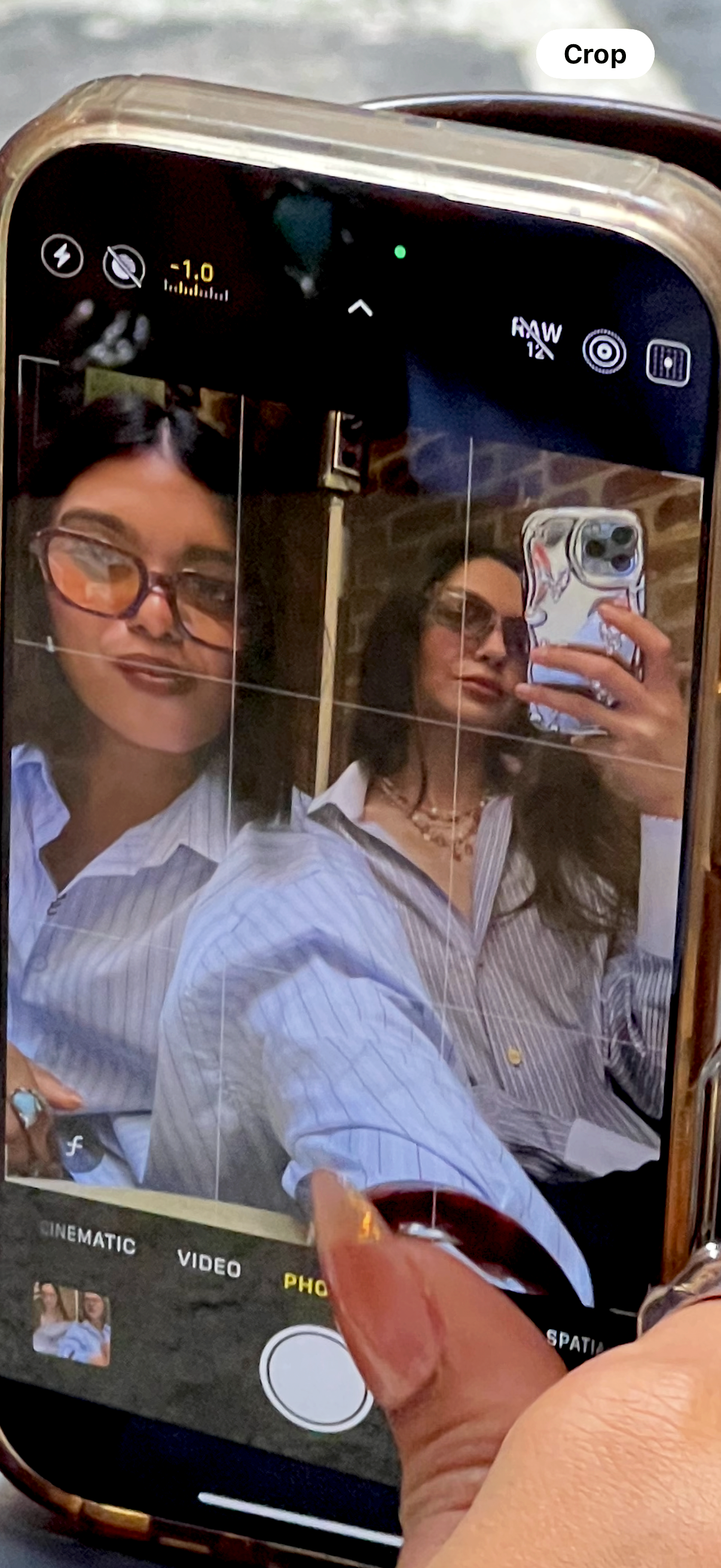 A person taking a mirror selfie with a smartphone, capturing two women in the reflection. The women are wearing glasses and striped shirts, posed indoors with a brick wall background.