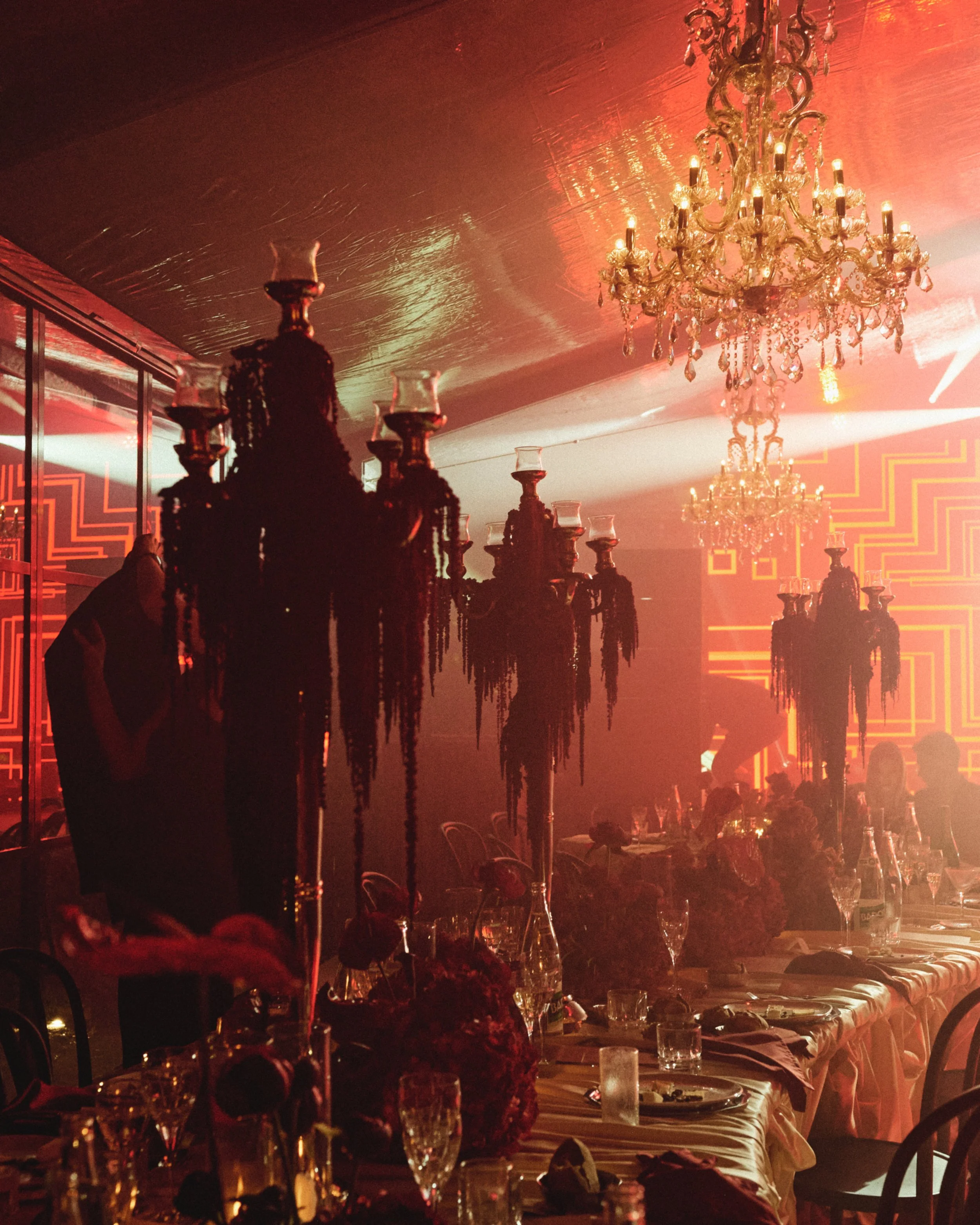 Elegant dining setup with long tables, chandeliers, candles, glassware, and tableware in a warmly lit venue.