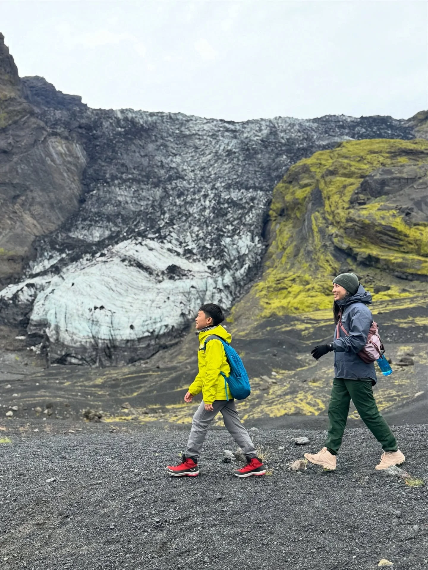 Two people hiking on a volcanic landscape with dark gravel and moss-covered rocks, and a glacier in the background.