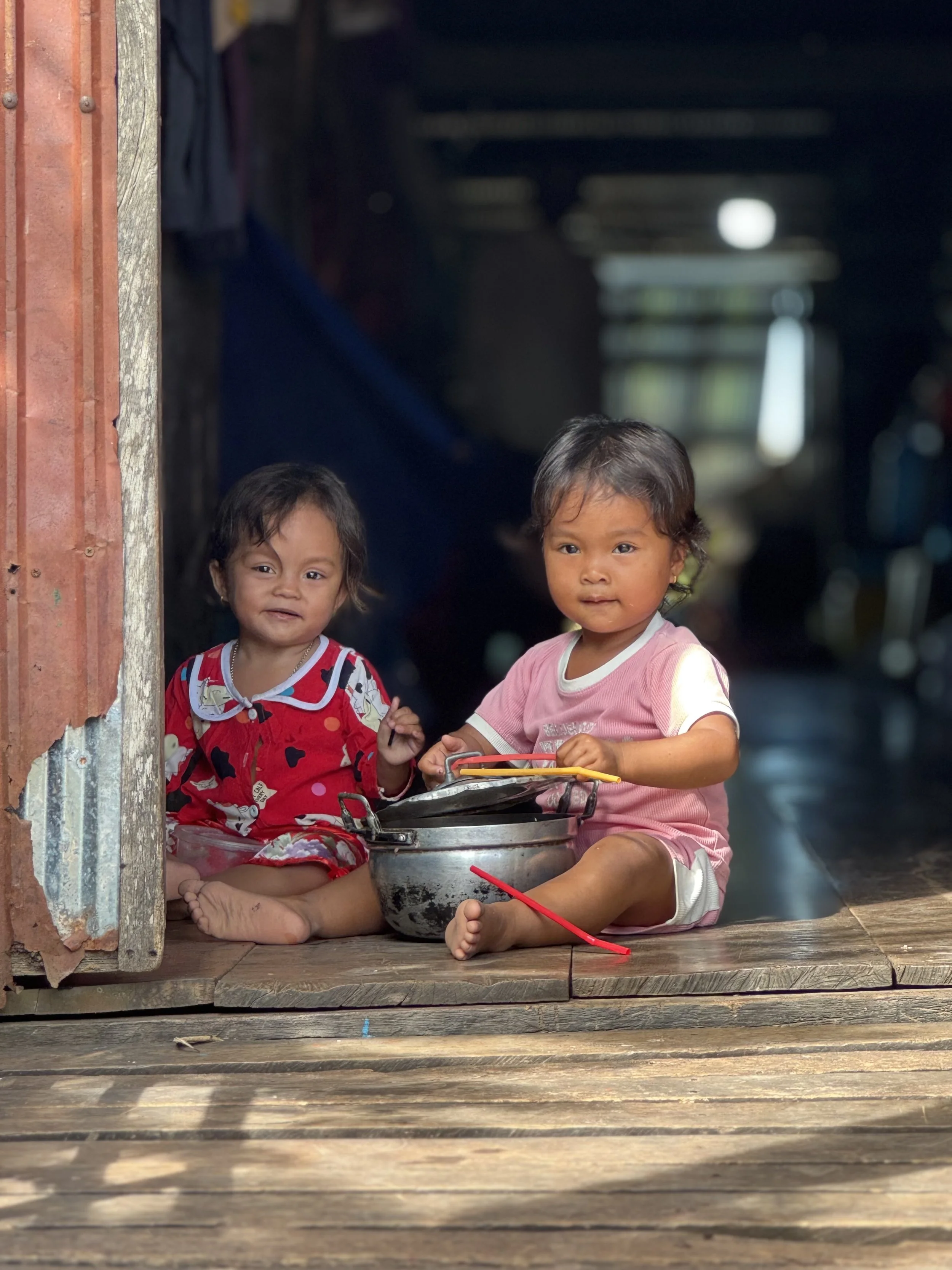 Two young children with dark hair and brown skin sitting on a wooden porch in front of a rustic building. One girl wears a pink shirt, and the other girl wears a colorful dress. They are near a pot and seem to be playing or resting.