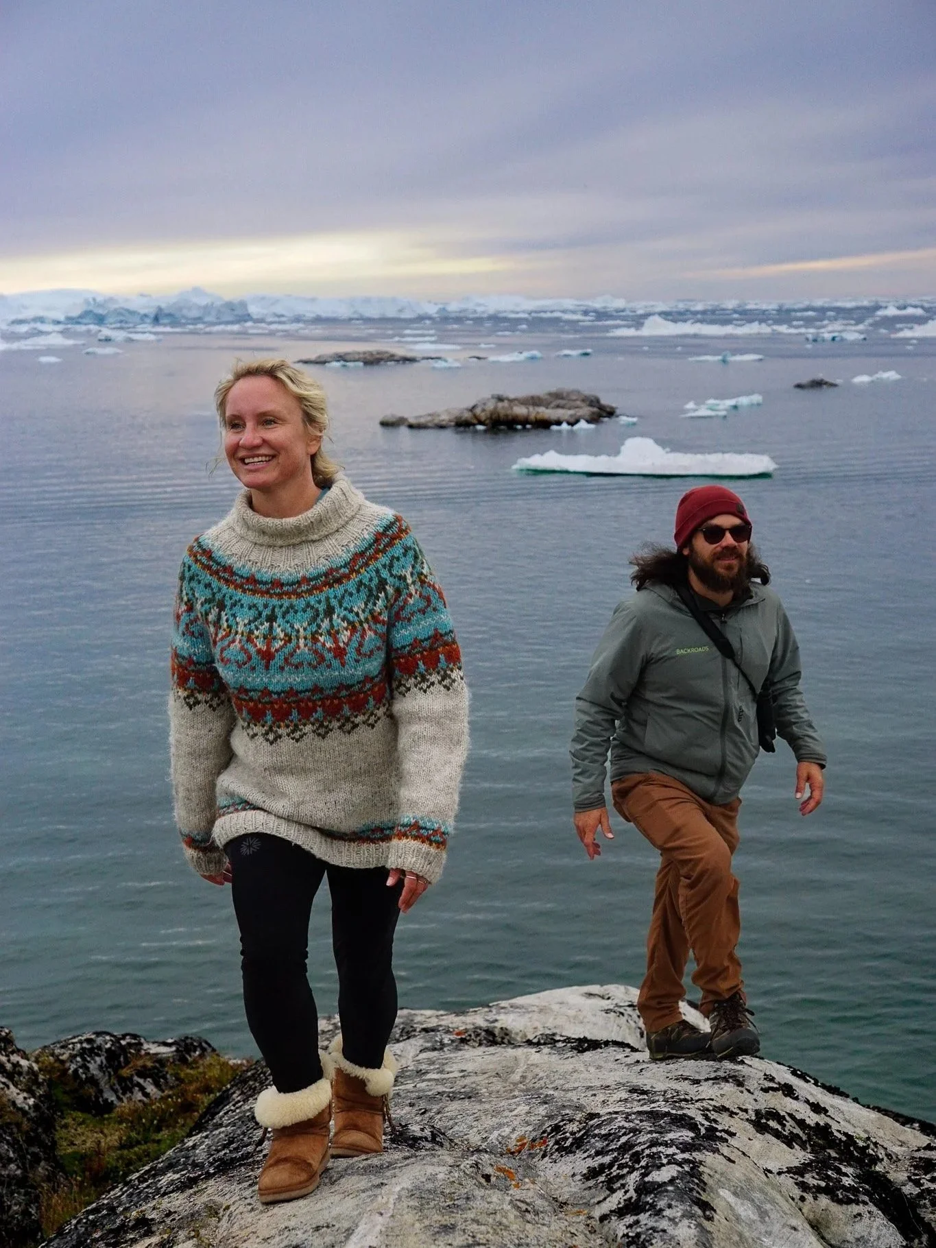 Two people standing on a rocky shoreline with icy water and icebergs in the background under a cloudy sky.