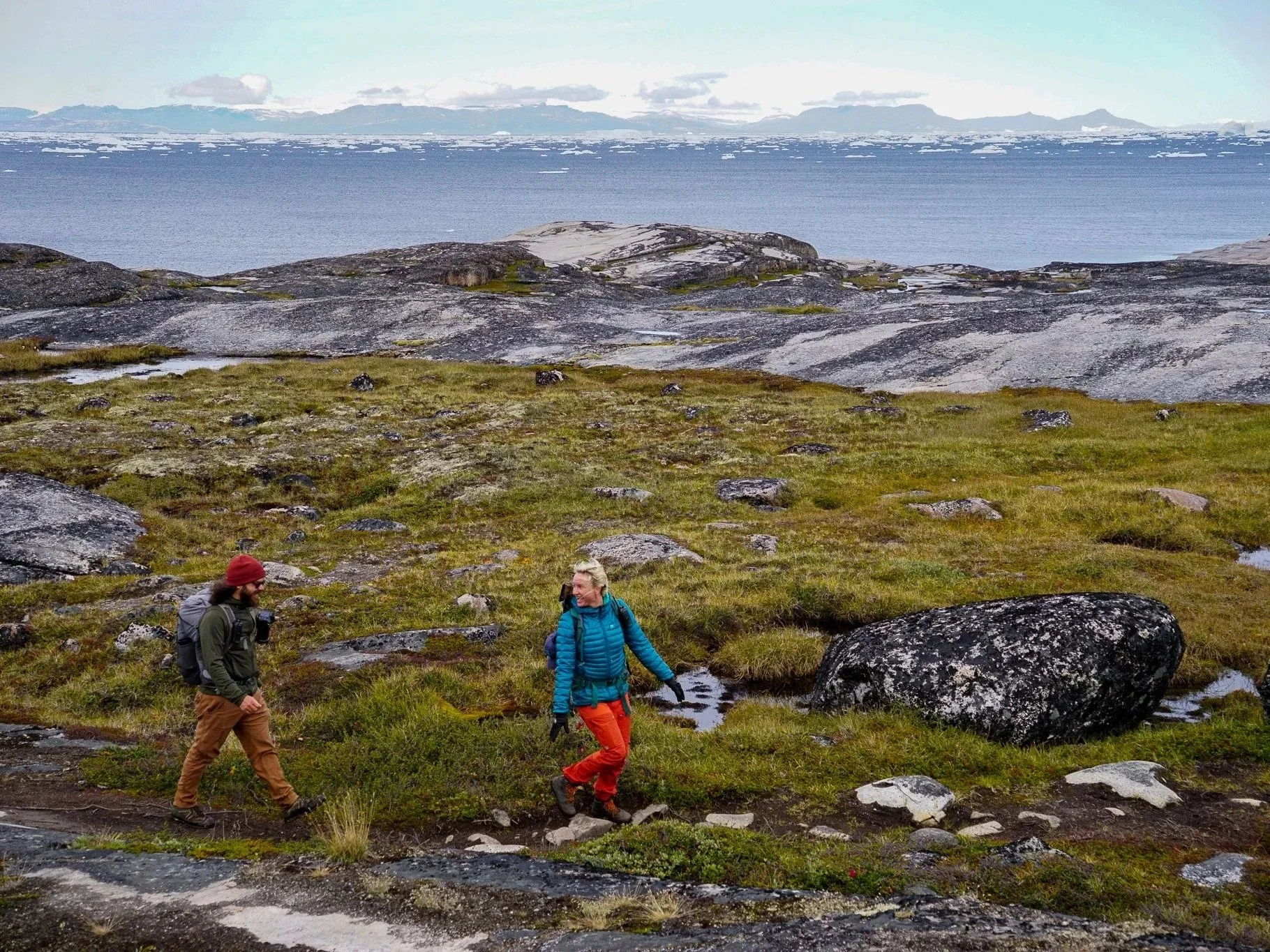 Two hikers walk along a rocky, grassy trail near a shoreline with large rocks, between green terrain and distant icy mountains across a body of water.