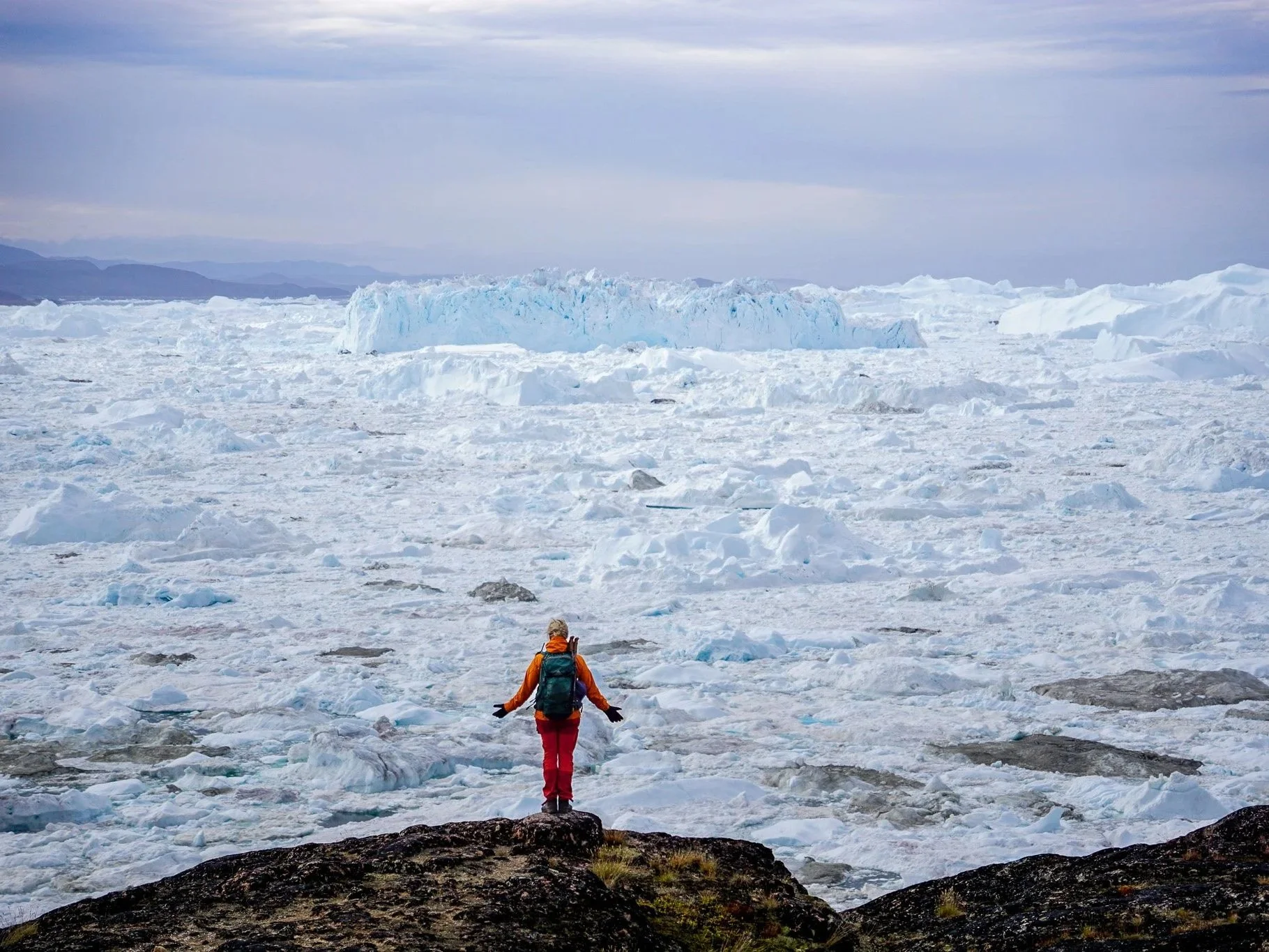 A person wearing an orange jacket, red pants, and a backpack stands on a rock in front of a vast icy landscape with ice and snow, looking towards a large glacier or ice formation under a cloudy sky.
