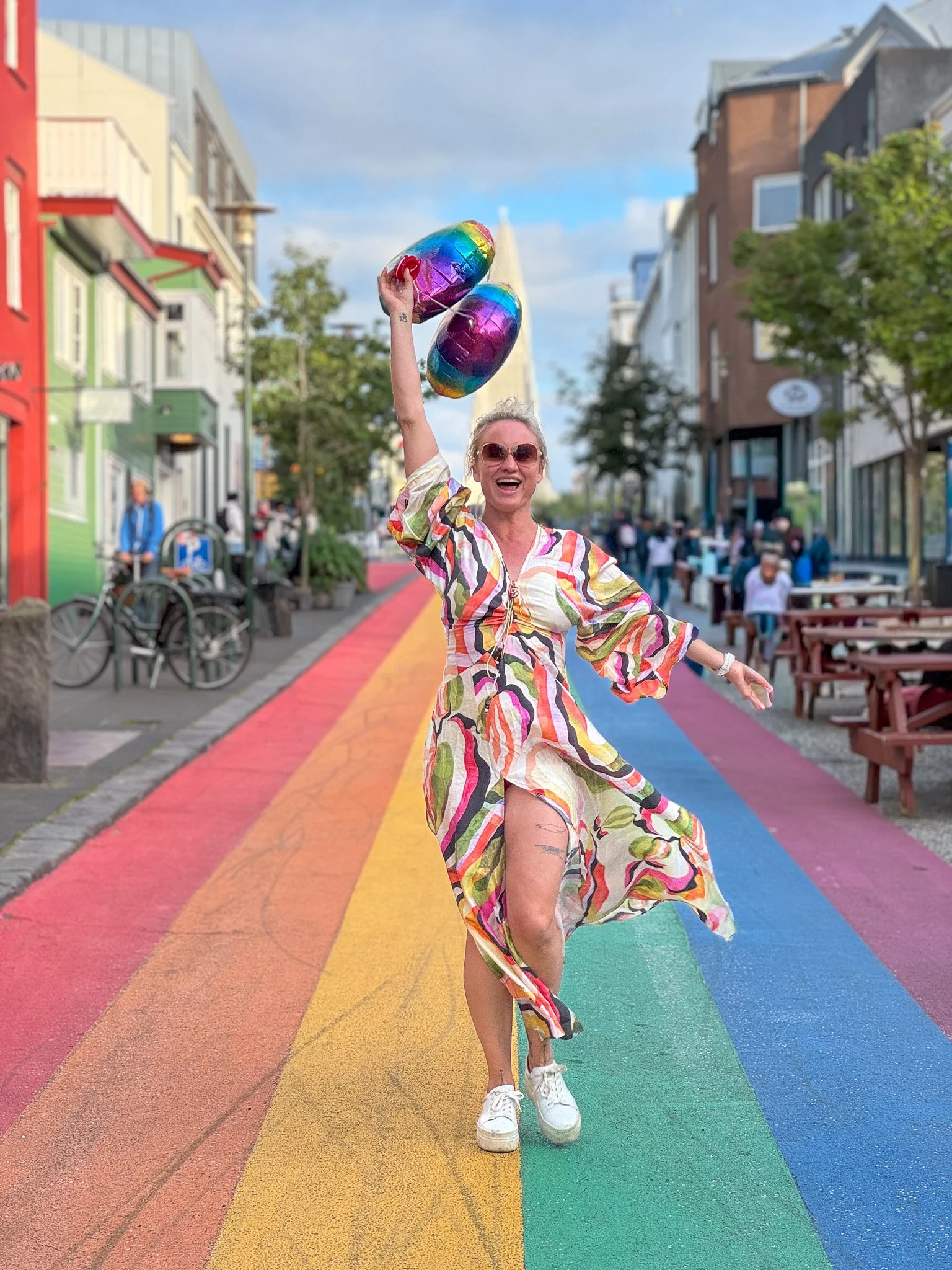 Woman in a colorful dress holding rainbow-colored balloons, standing on a rainbow-painted crosswalk on a city street with buildings, trees, and people in the background.
