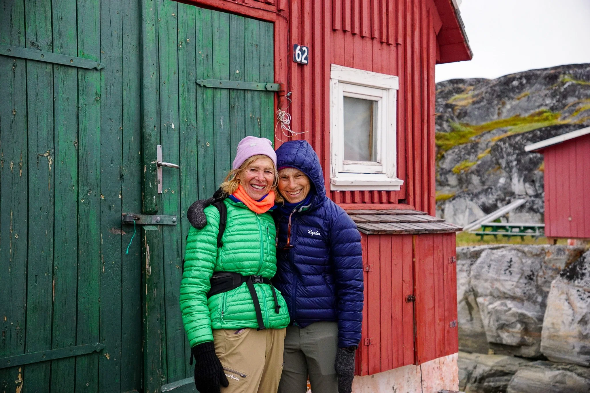 Two women smiling and posing outdoors in front of a green and red wooden building. They are wearing winter jackets and hats.