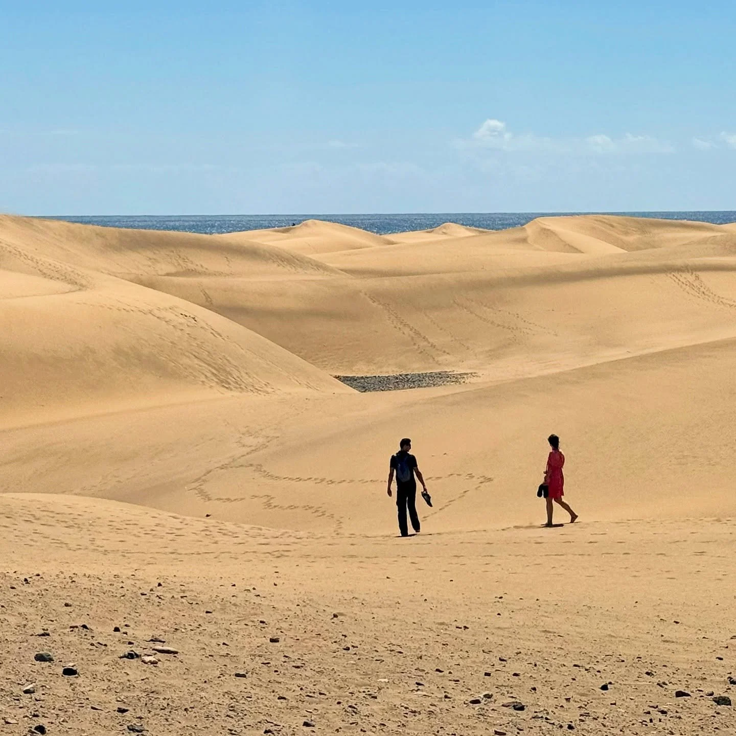 Two people walking on sandy dunes with the ocean and blue sky in the background.