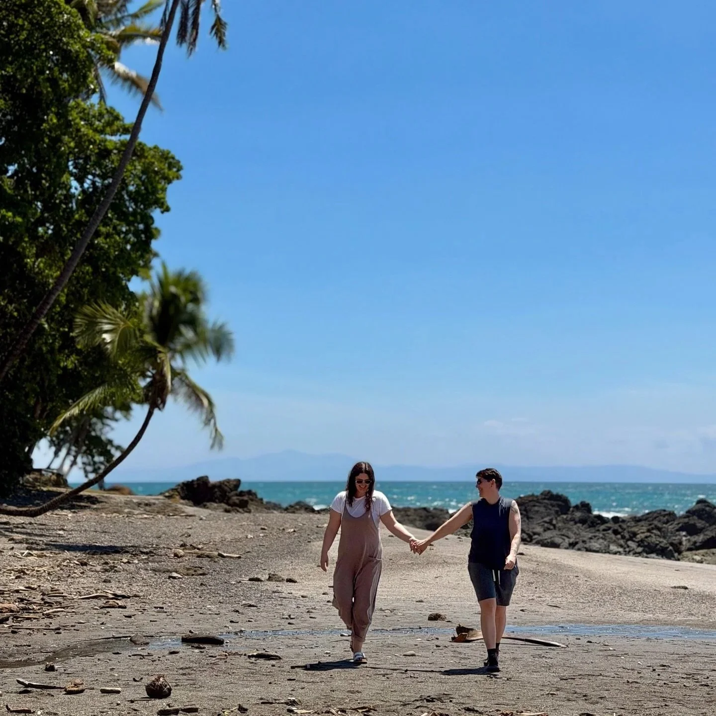 A young couple walking hand in hand on a sandy beach with rocks, palm trees, and the ocean in the background on a sunny day.