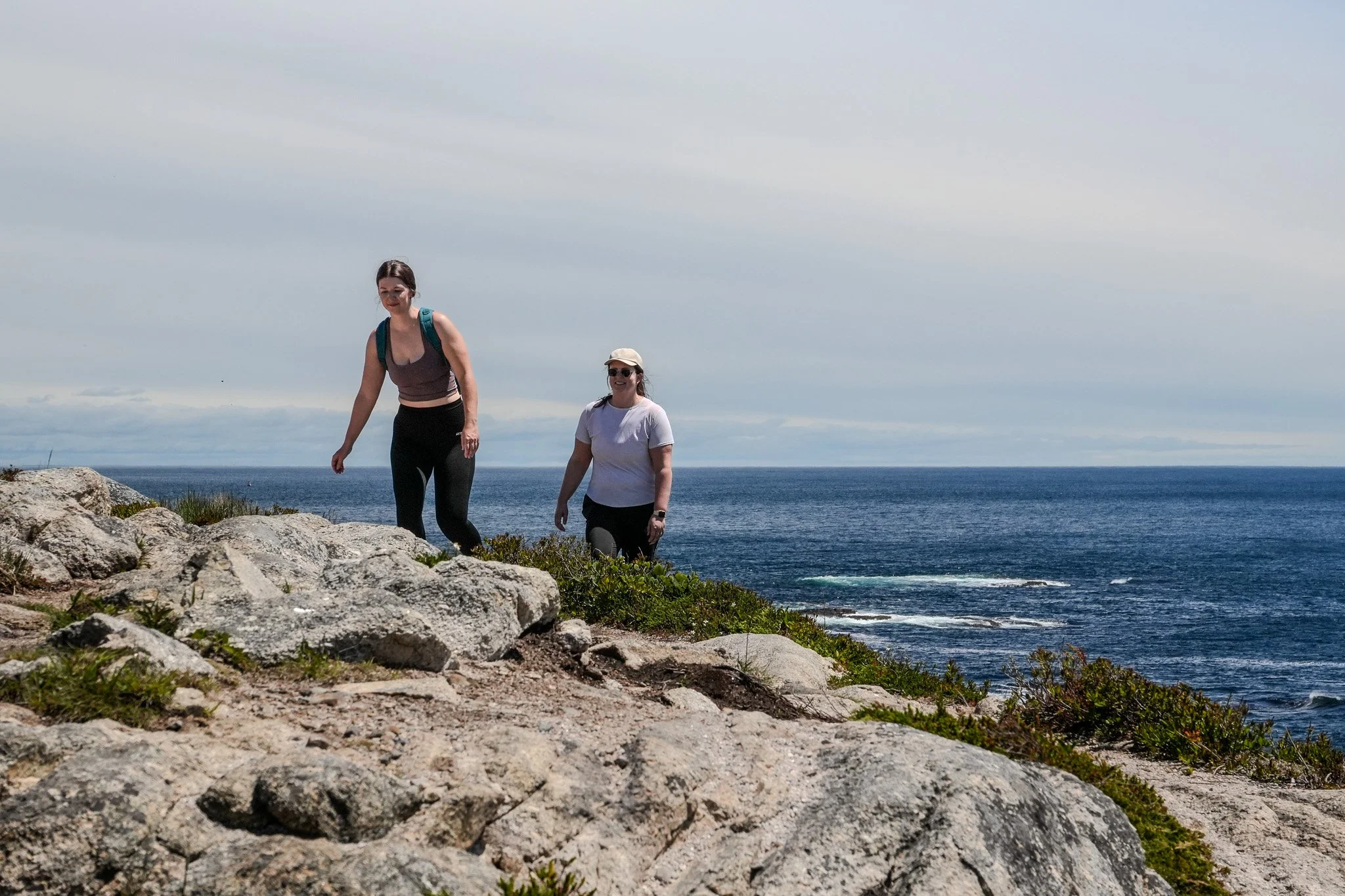 Two women hiking on rocky terrain near the ocean, with cloudy sky overhead.