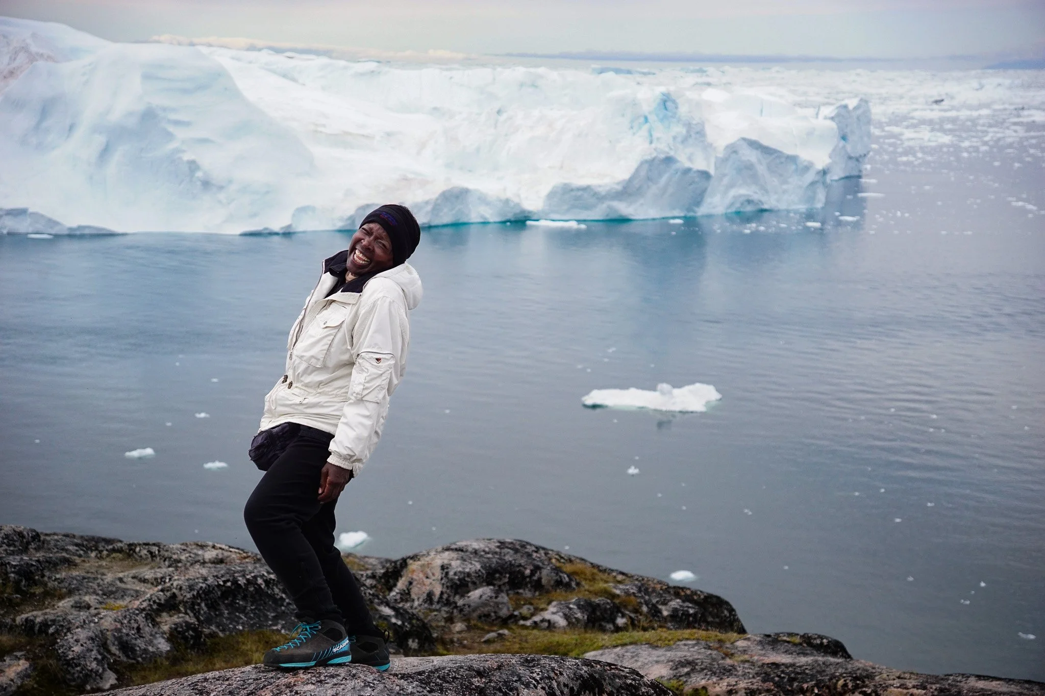 A person standing on rocks near the water with large icebergs and glaciers in the background.