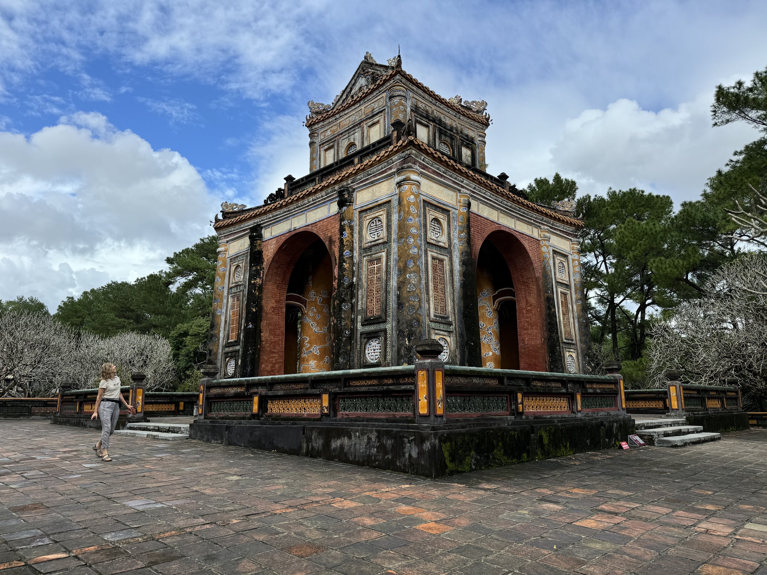 A historical stone and brick pavilion with ornate decorations and a tiled roof, set in a park with trees and a cloudy sky in the background.