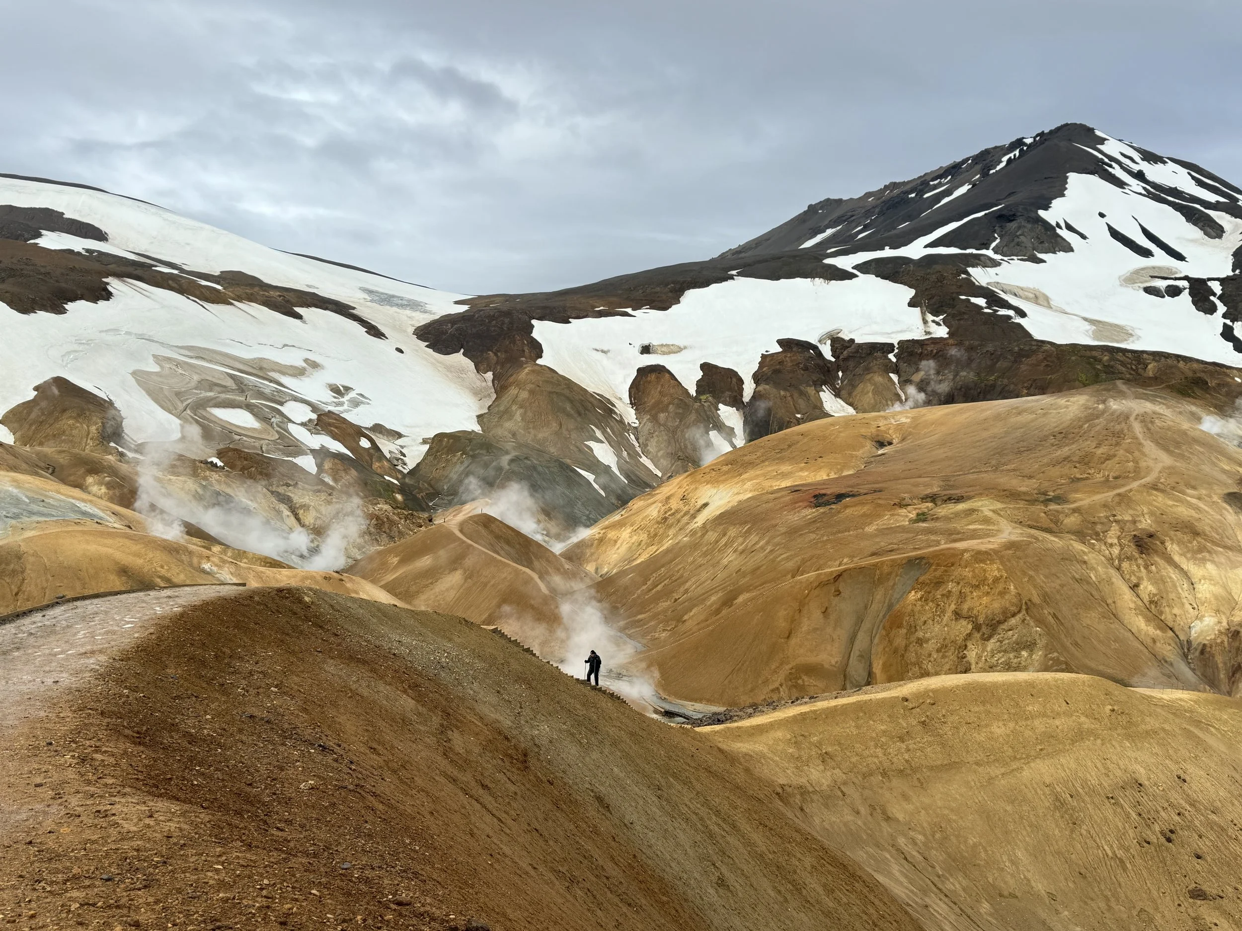 A landscape featuring a mountain with snow patches, colorful geothermal ground, and a person in the foreground.