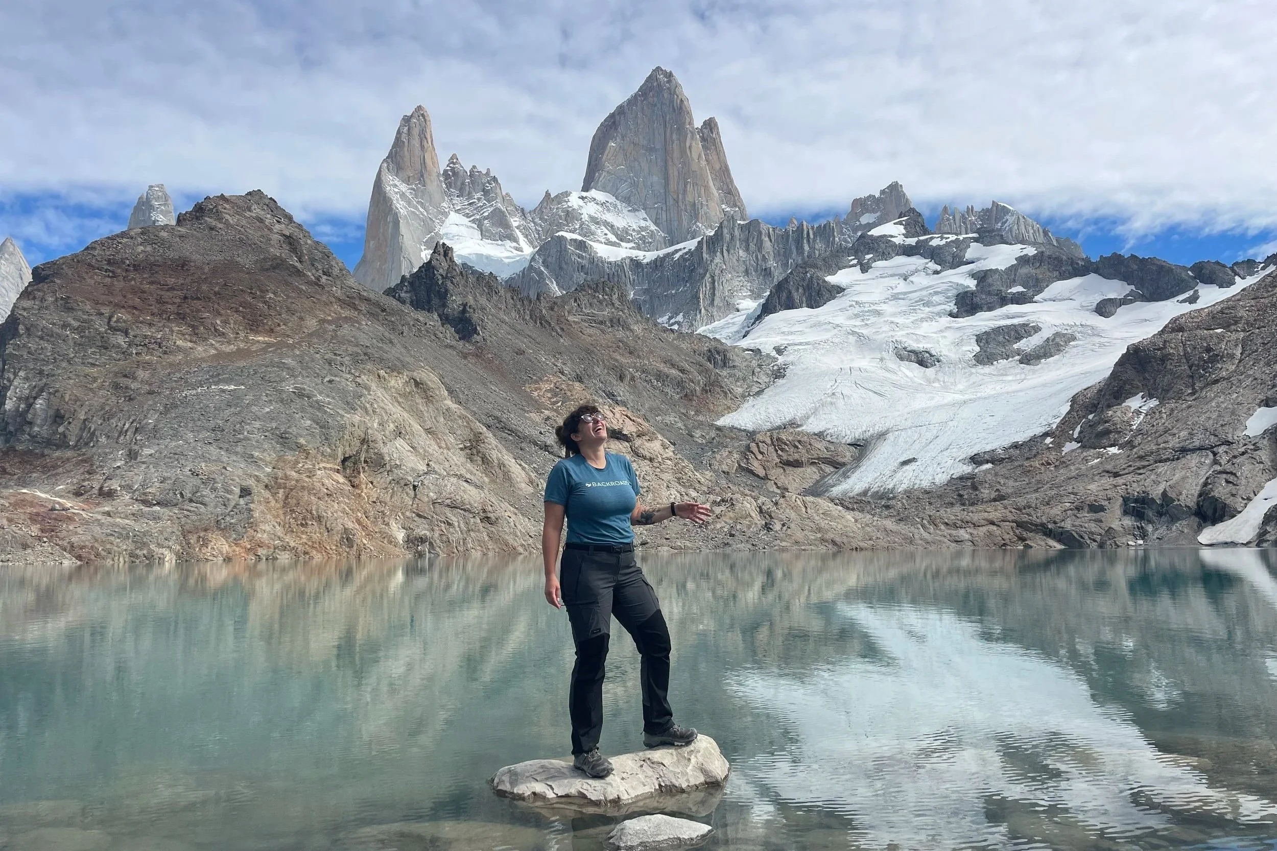 A woman standing on a rock in a lake with mountains, snow, and glaciers in the background.