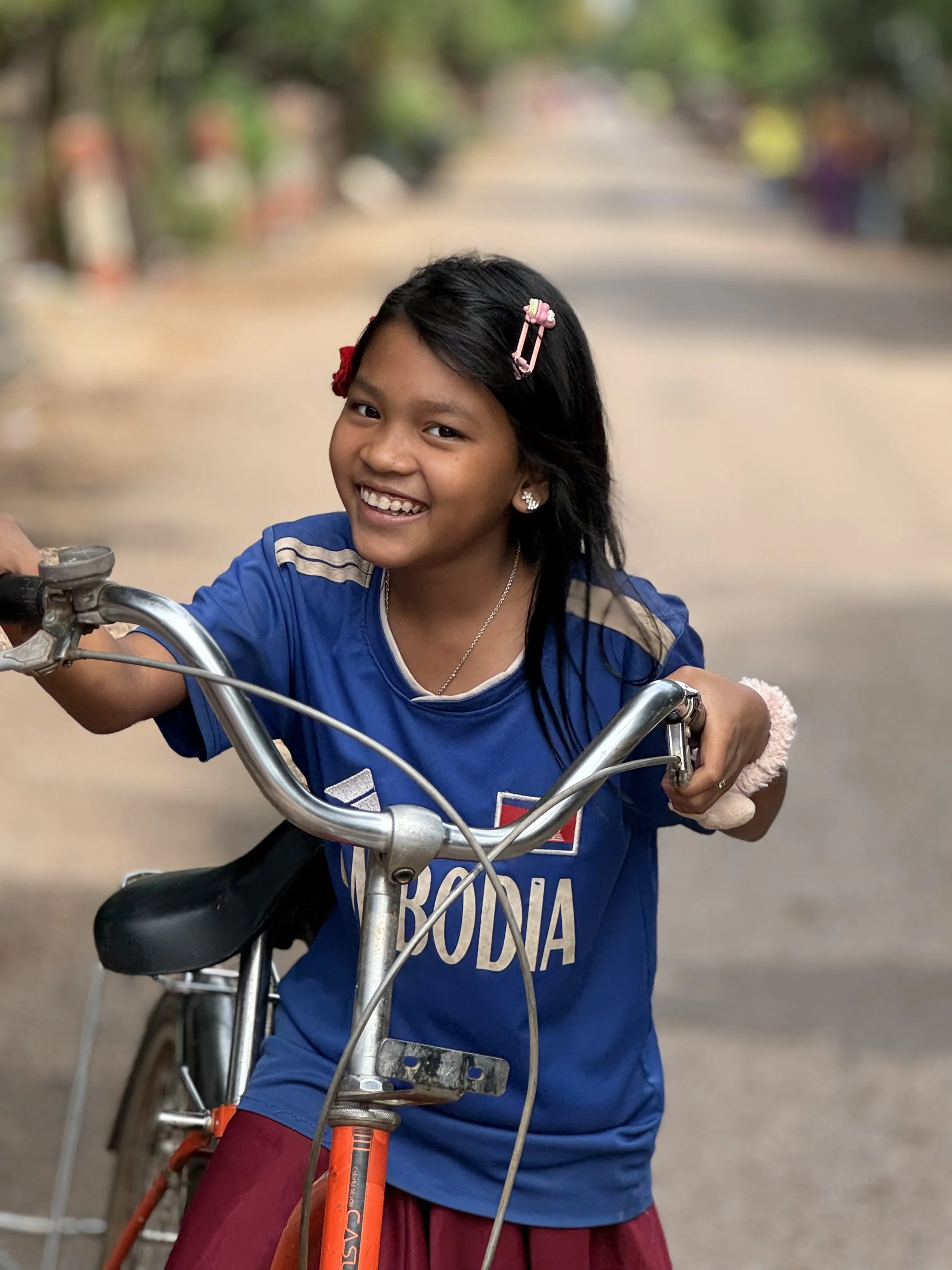 A young girl with dark hair, smiling, riding a bicycle on a blurry outdoor street with colorful houses and trees in the background.