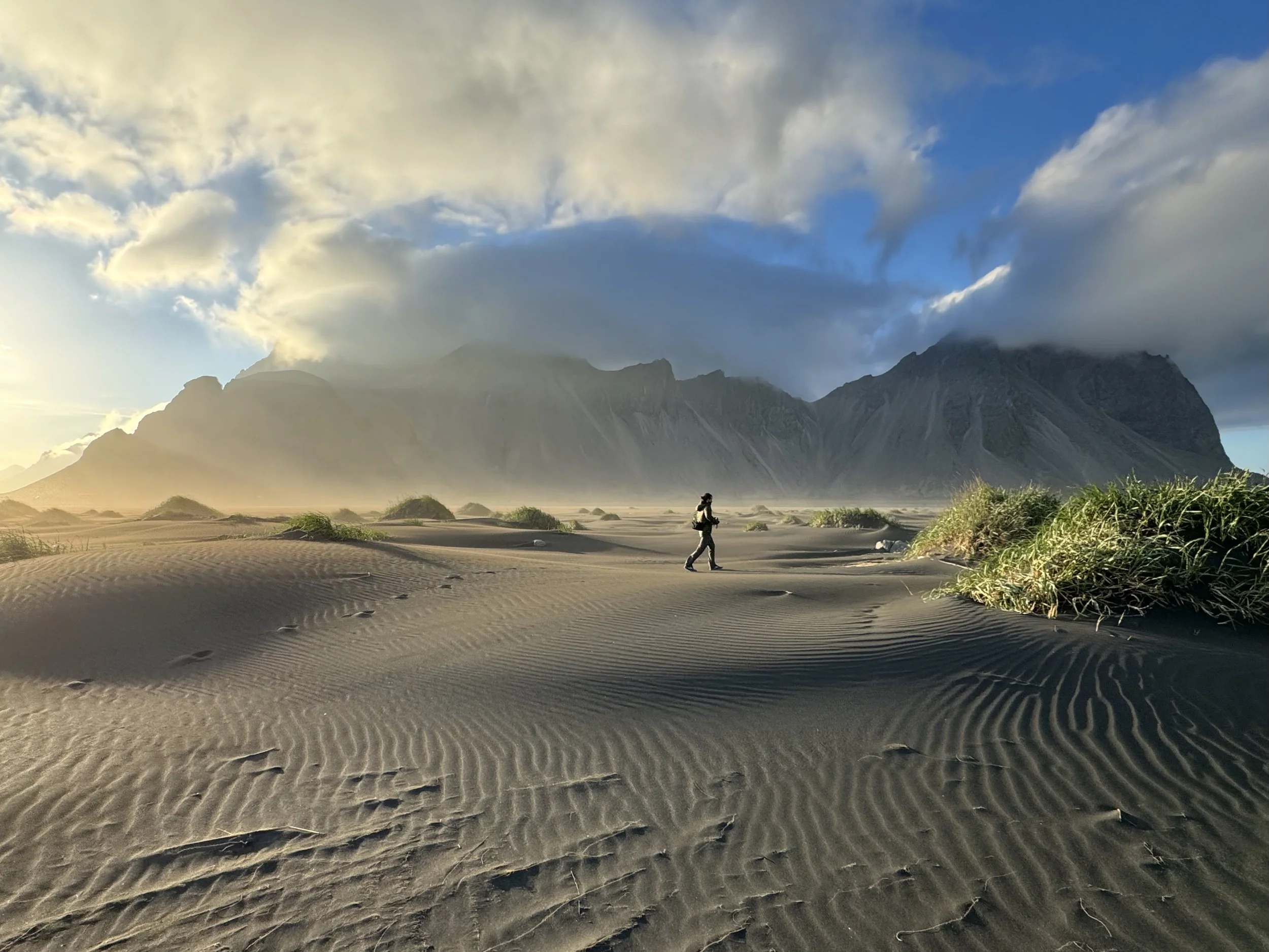 A person walking across a sandy desert with mountain peaks and clouds in the sky in the background.