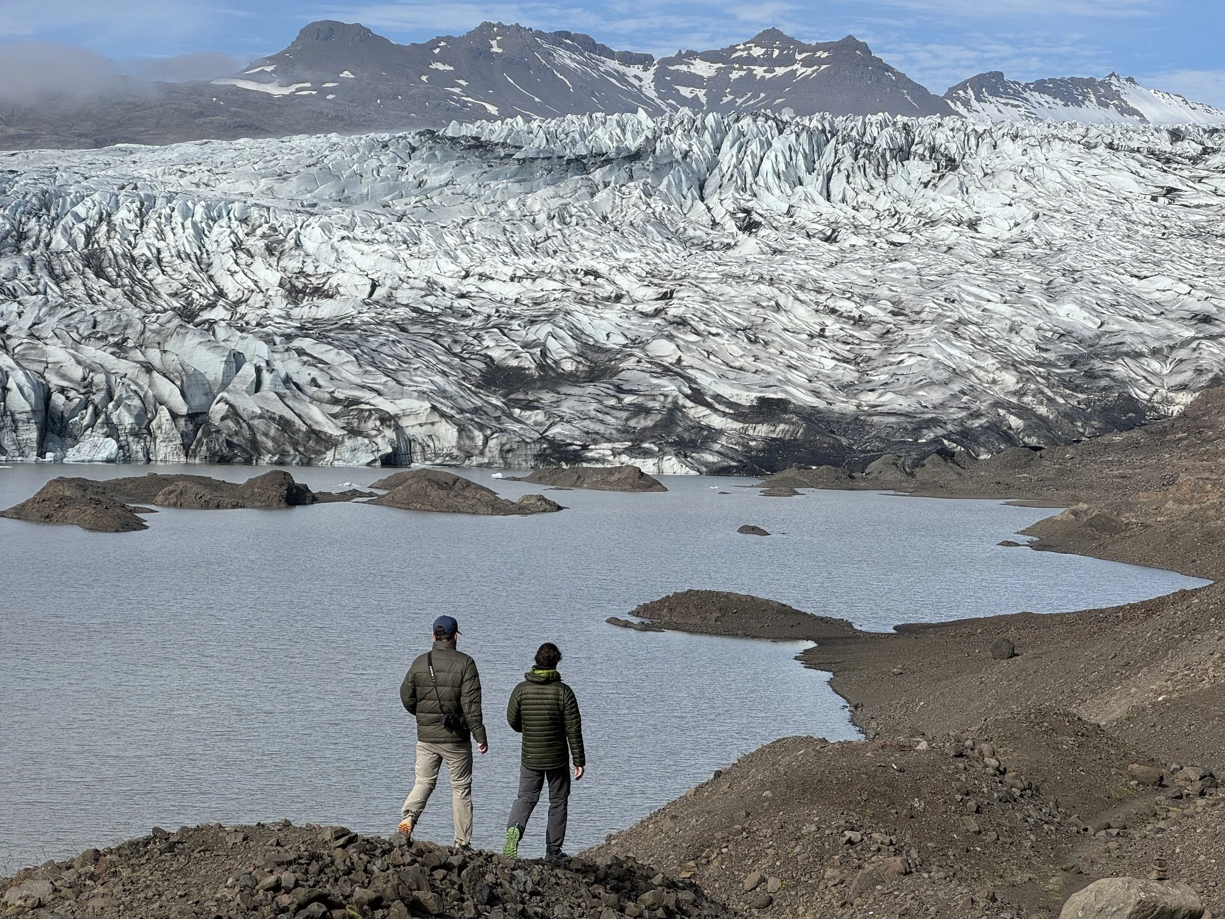 Two people in jackets walking along a rocky shore towards a glacier with mountains in the background.
