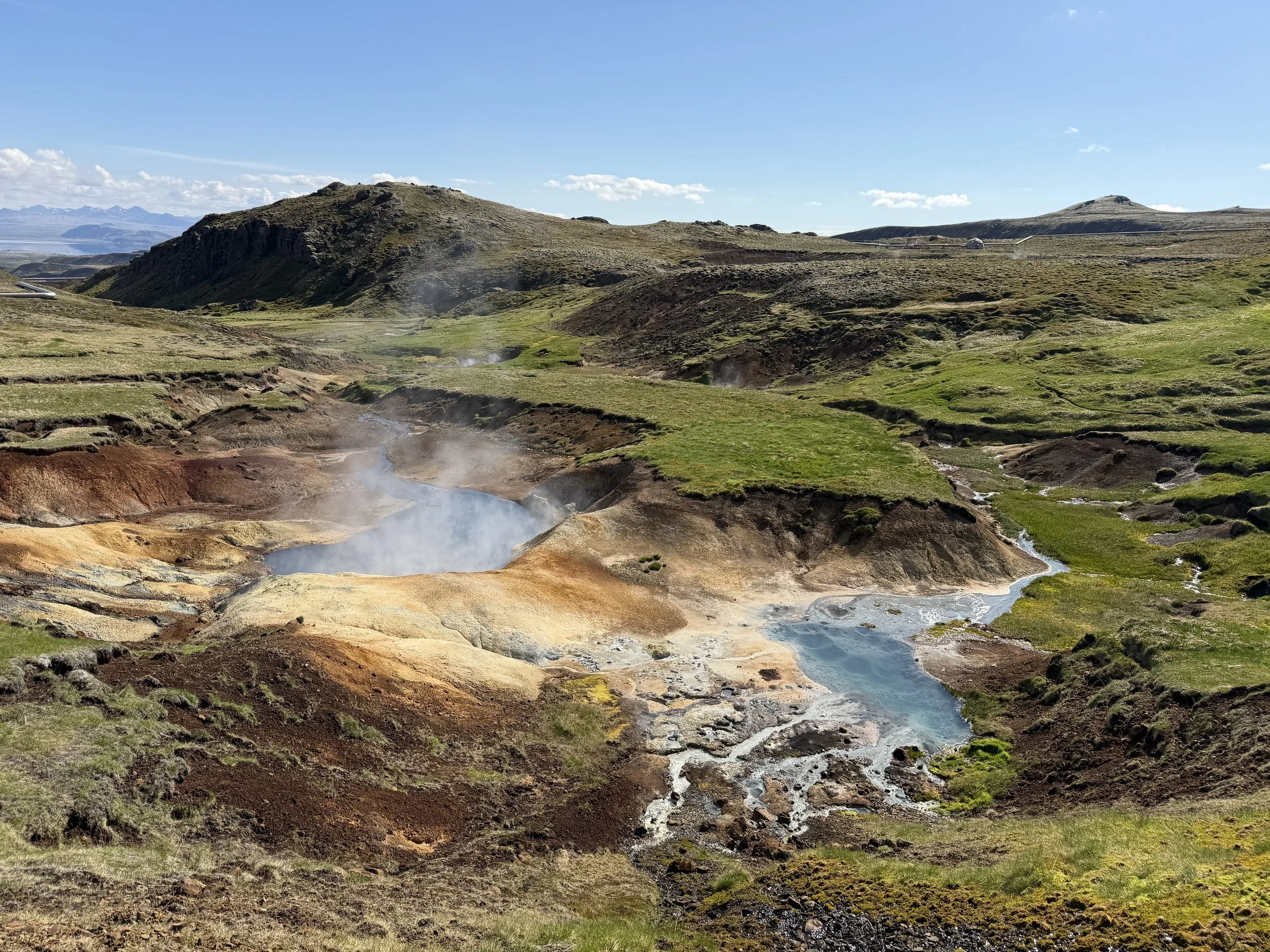 A scenic landscape with steaming geothermal hot springs, green grassy patches, and brown rocky hills under a clear blue sky.