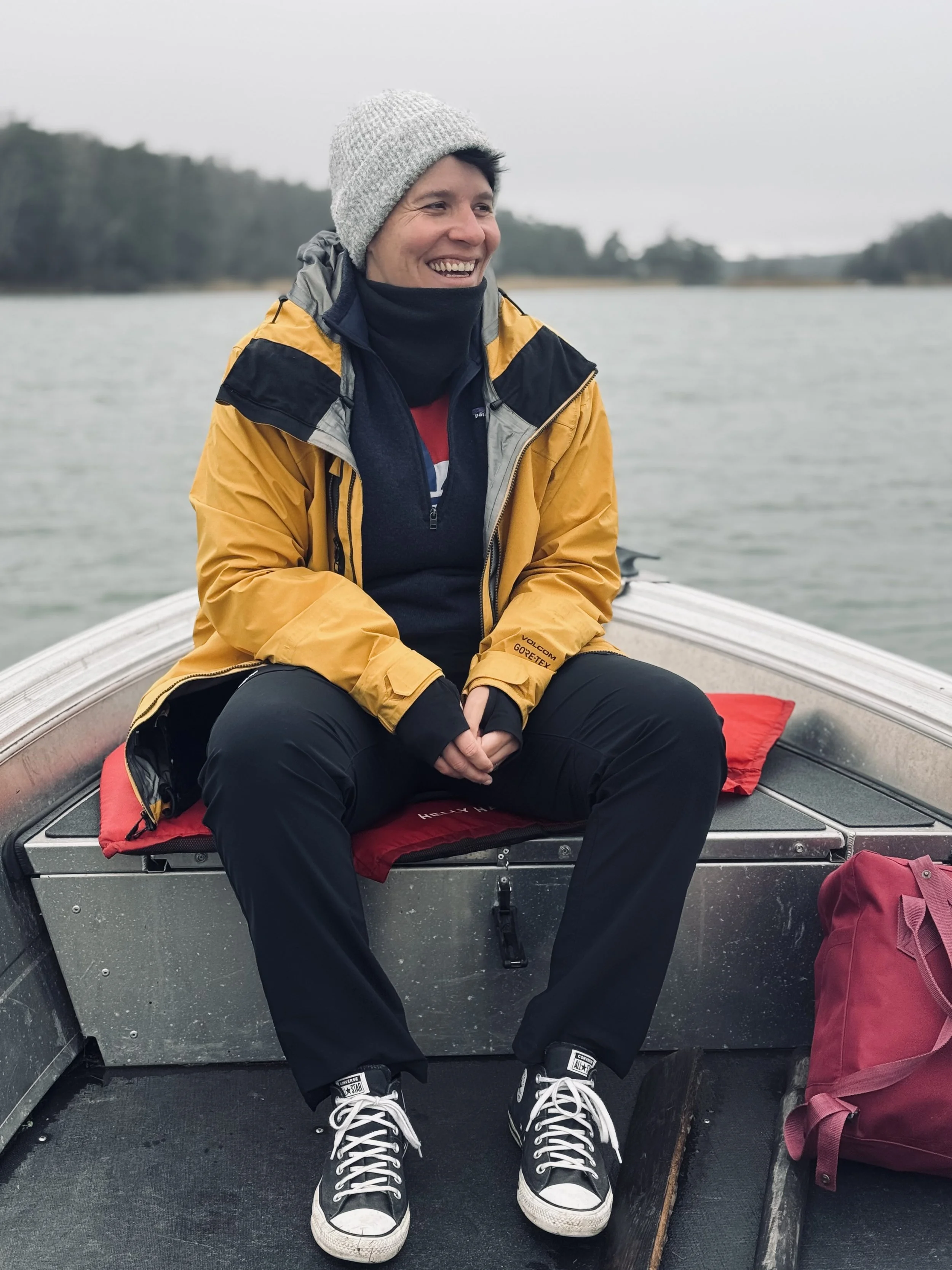 A person sitting on a boat wearing a yellow and black jacket, black pants, and black and white sneakers, with trees and water in the background.