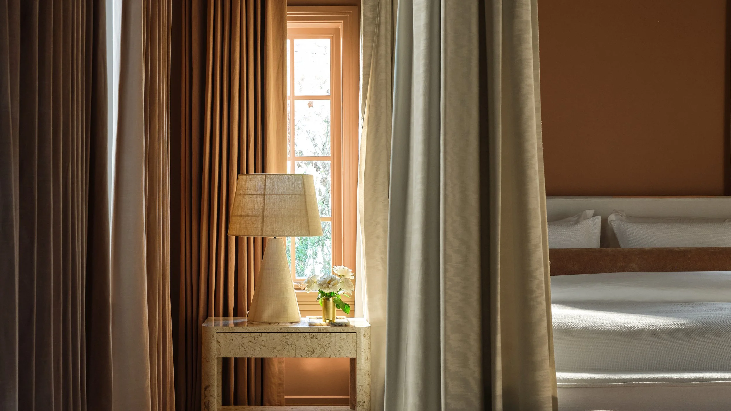A window with beige and brown curtains, a side table with a beige lamp and a small vase of white flowers, and a bed with white bedding partially visible behind the curtains.