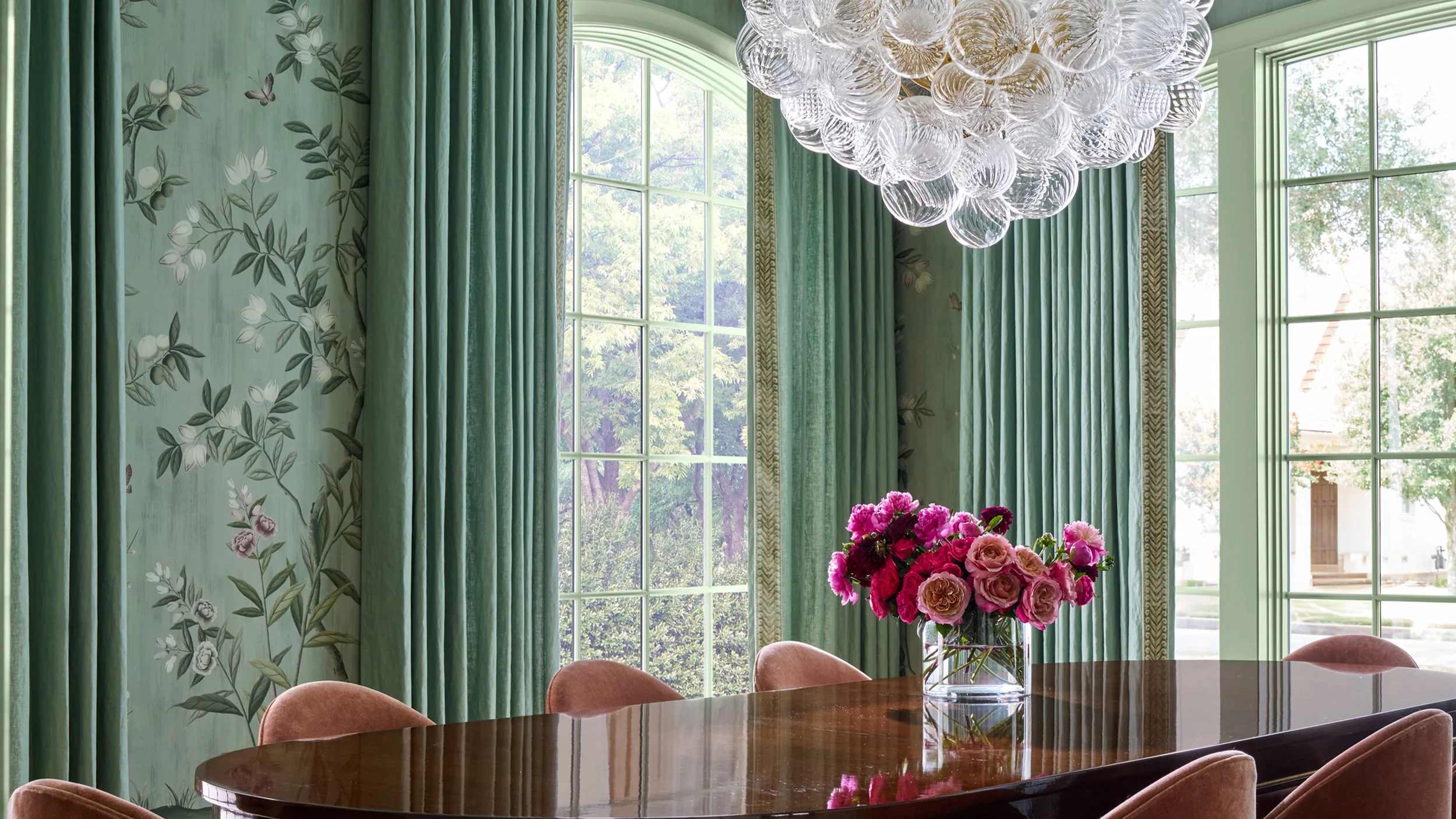 A dining room with green floral wallpaper, large windows with green curtains, a chandelier made of glass orbs, a vase of pink flowers on a wooden table, and pink upholstered chairs.