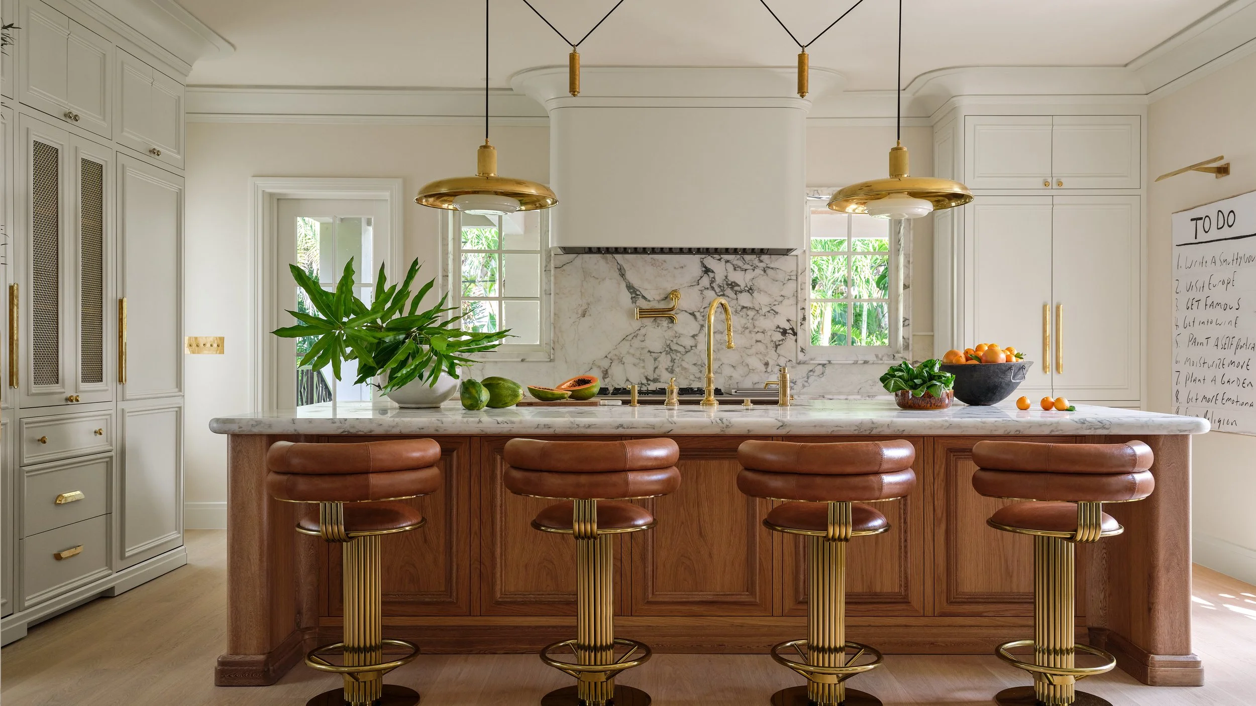 A modern kitchen with white cabinets, a marble backsplash, and a marble island. The island has a wooden base and is surrounded by five brown leather bar stools with gold accents. Two gold pendant lights hang above the island. There is a green plant, a bowl of oranges, and some cut papayas on the island.