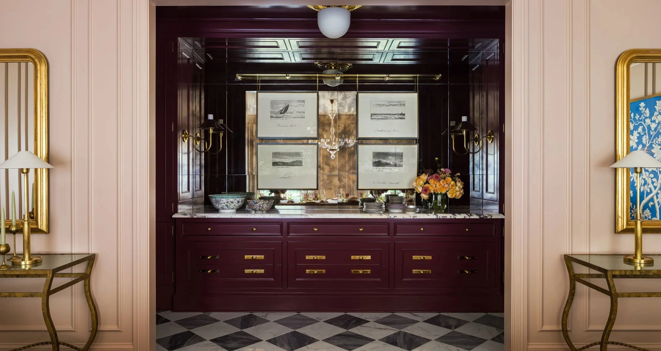 Interior view of a dining area with a built-in burgundy cabinet, framed black and white artwork, a chandelier, flower vase with pink and yellow flowers, and decorative bowls, flanked by gold wall sconces and gold-framed mirrors.