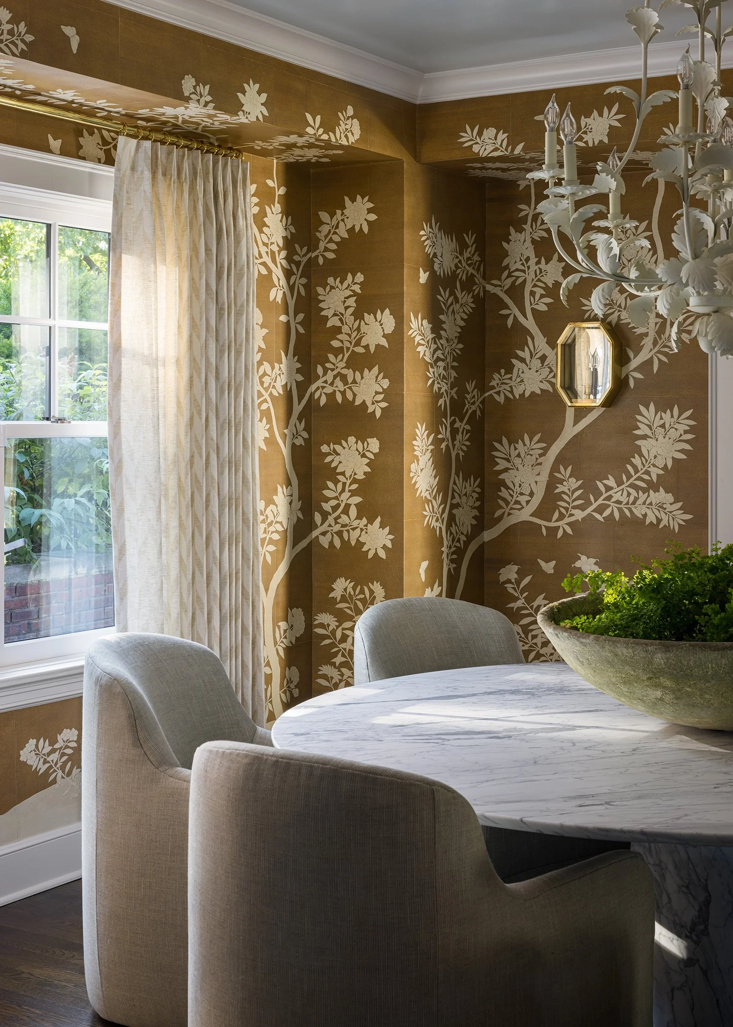 A dining room with beige upholstered chairs around a white marble table, a large potted plant, a window with cream-colored curtains, floral and butterfly patterned wallpaper in gold and white, a white chandelier, and a small mirror on the wall.