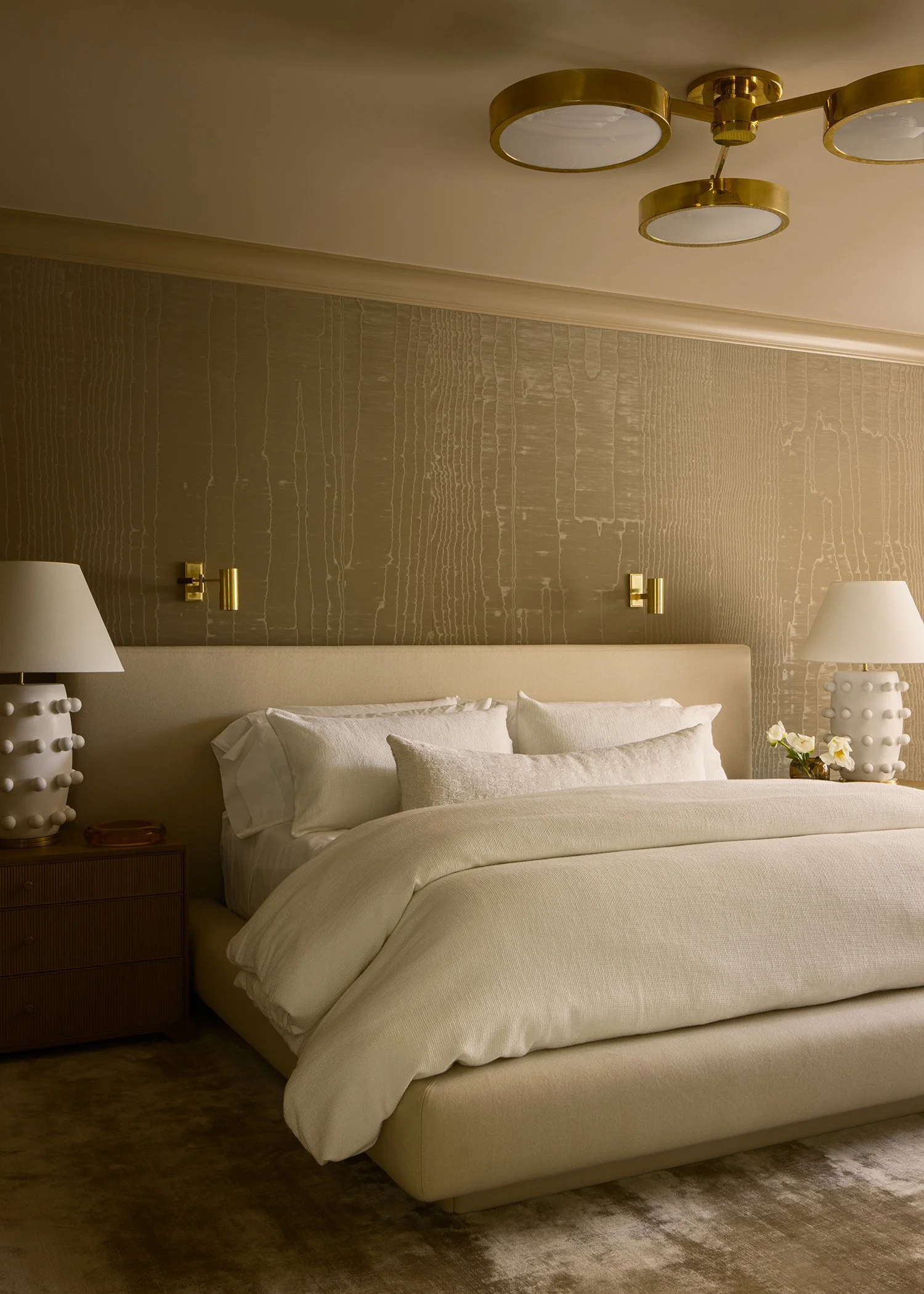 A bedroom with a large bed covered in white linens, two matching white nightstands with white lamps with decorative bases, a textured beige wallpaper behind the bed, and a modern gold ceiling light fixture.