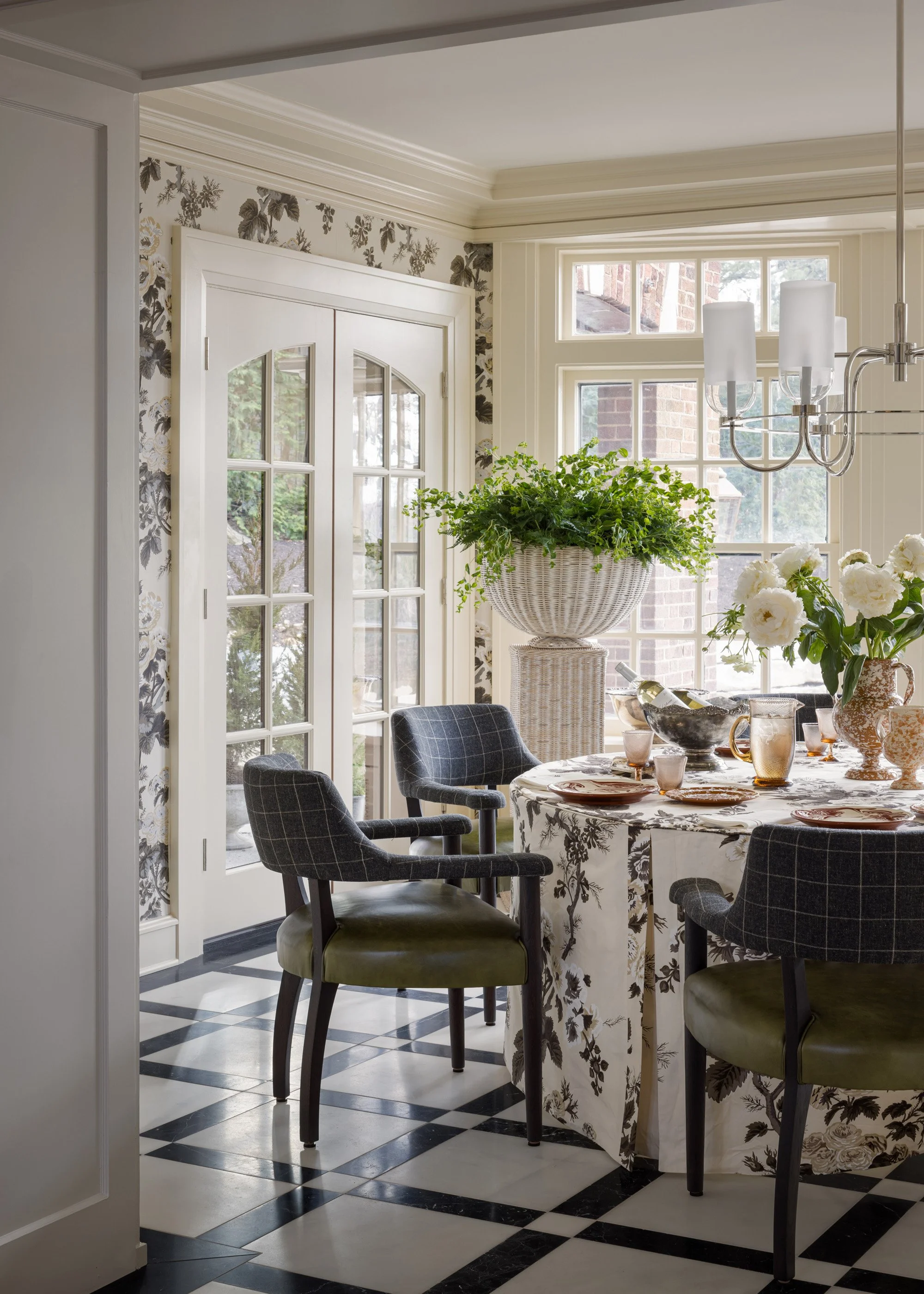 A dining room with a round table covered with a floral tablecloth, surrounded by black plaid chairs with green cushions, large windows, a chandelier with white shades, green potted plants, and a black and white checkered floor.