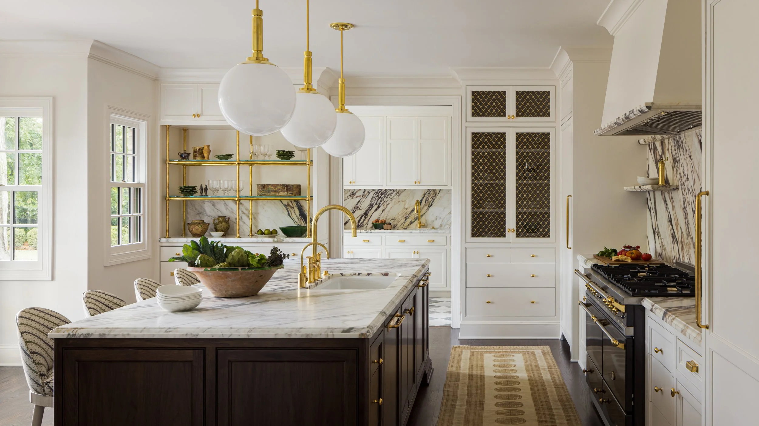 A bright, elegant kitchen with white cabinetry, marble countertops, and gold accents. It features a large island with a dark wood base, a marble top, and gold fixtures. Hanging from the ceiling are four large white spherical pendant lights. There are