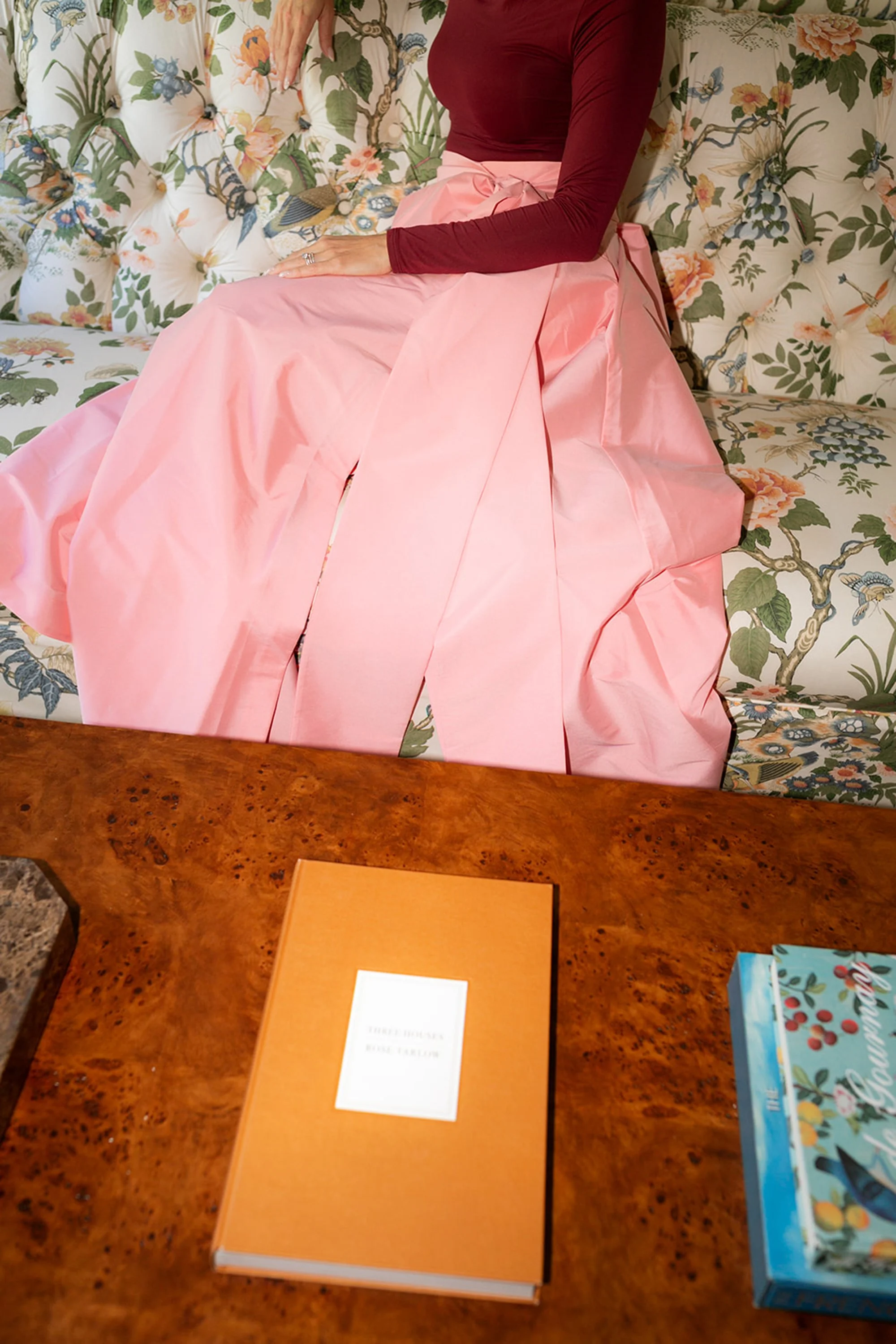 A woman wearing a burgundy long sleeve shirt and a long pink skirt sitting on a floral patterned couch with a hand resting on her lap. There is a wooden table in front of her with an orange book and other colorful books around.