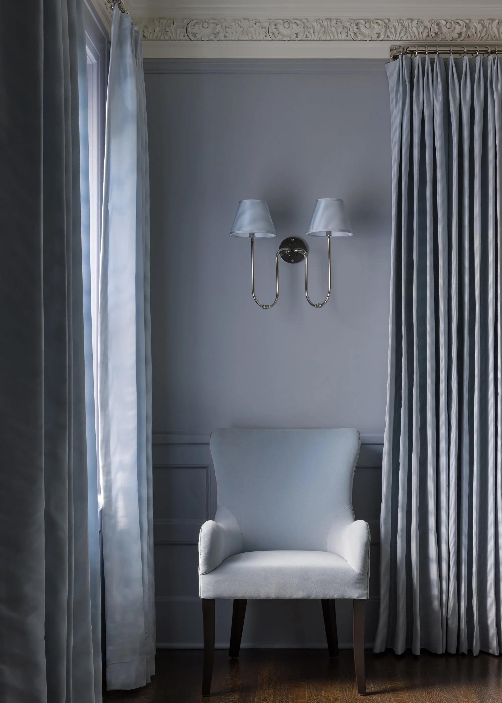 A white upholstered armchair with wooden legs positioned in front of a grey wall with decorative molding. Two wall-mounted lamps with white shades are above the armchair. Light curtains are on both sides of the chair, covering windows.