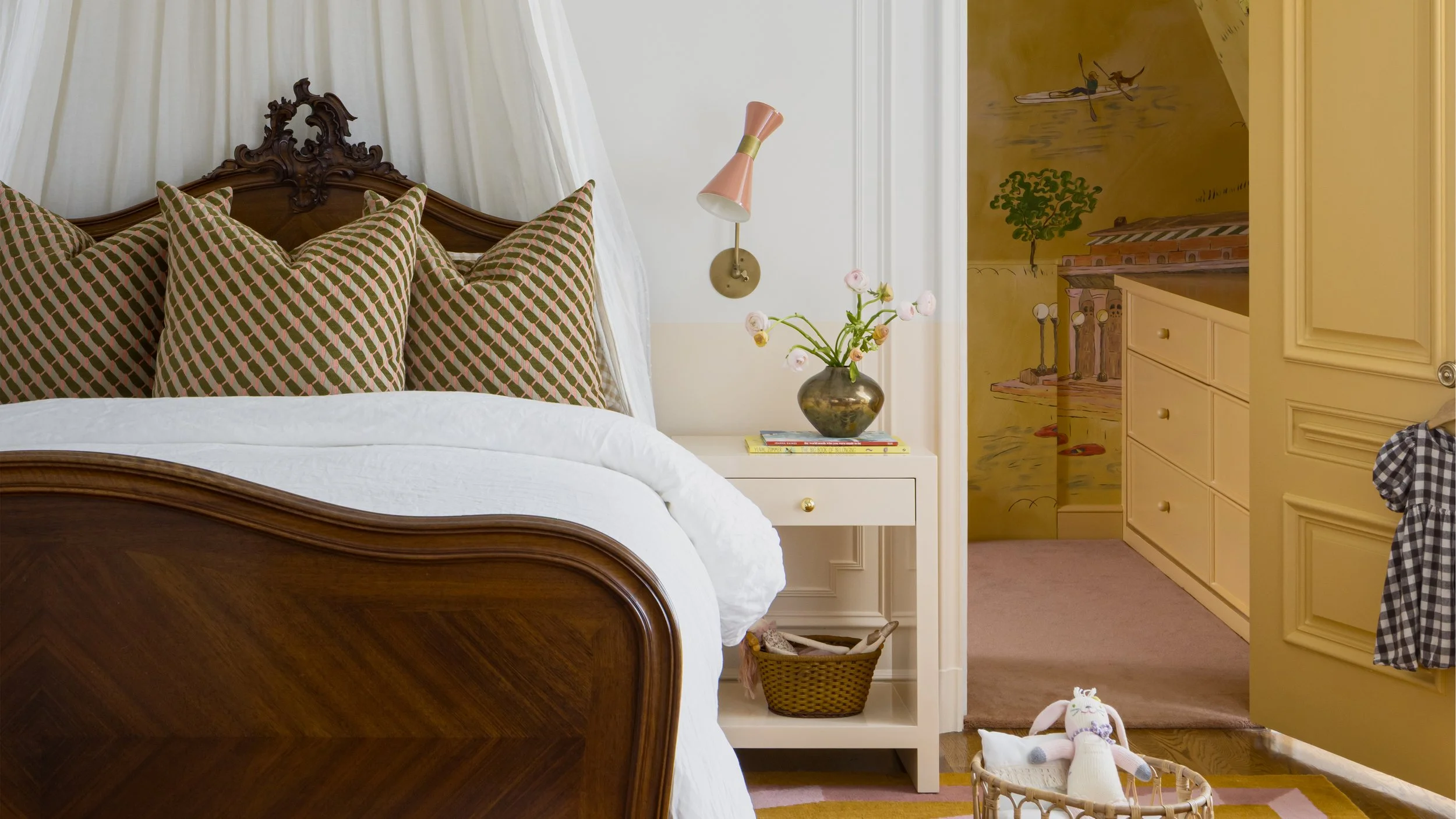 A bedroom with a wooden bed frame, pillows with geometric patterns, a white nightstand with books and a vase of flowers, and a pink wall-mounted lamp. There's a yellow door and a mural of a person paddling a canoe on the wall.