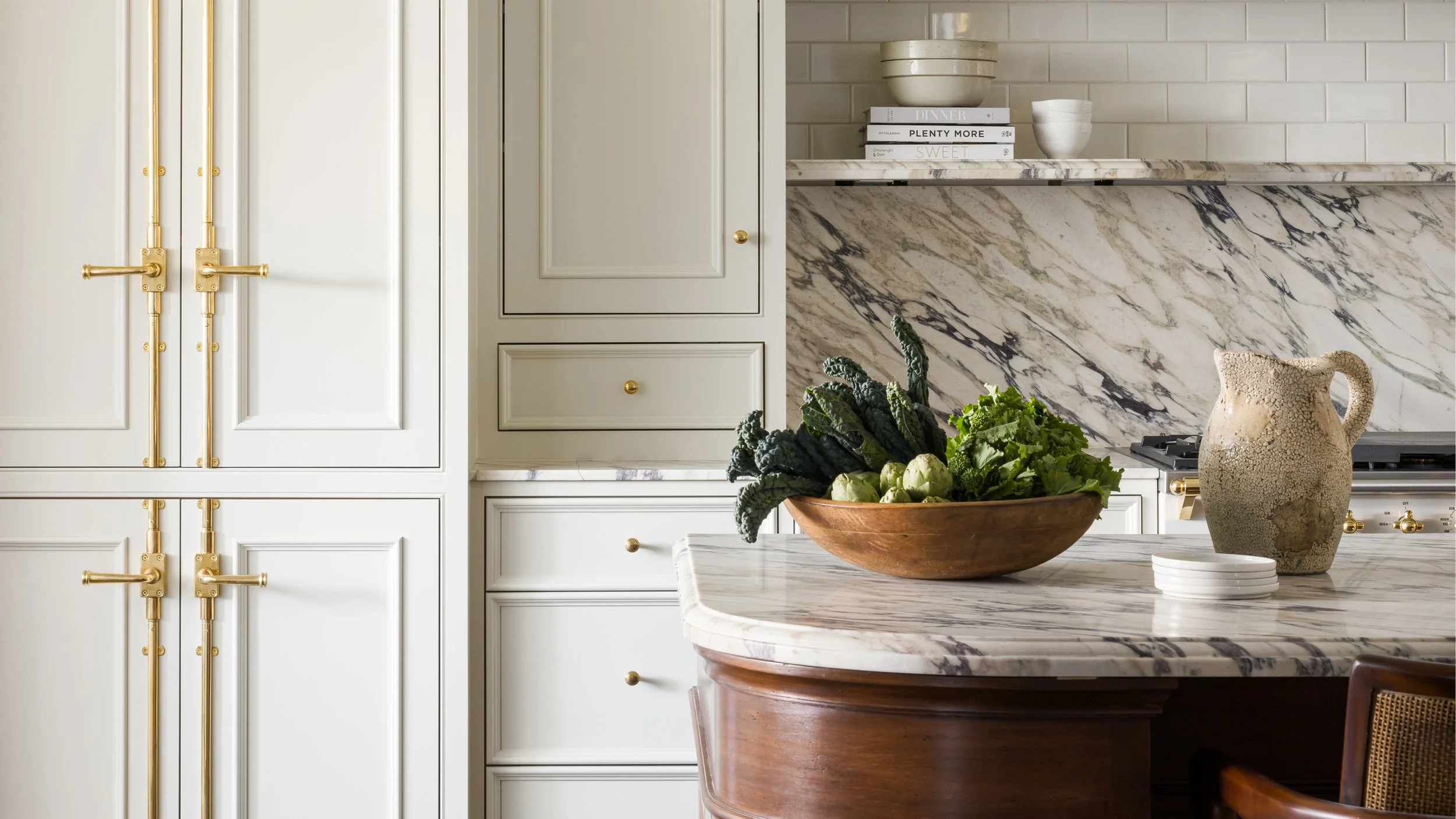 A kitchen with white cabinets, gold handles, and a marble countertop. There is a bowl of vegetables, a clay pitcher, and a stack of small white plates on the countertop, with a backsplash made of white subway tiles.