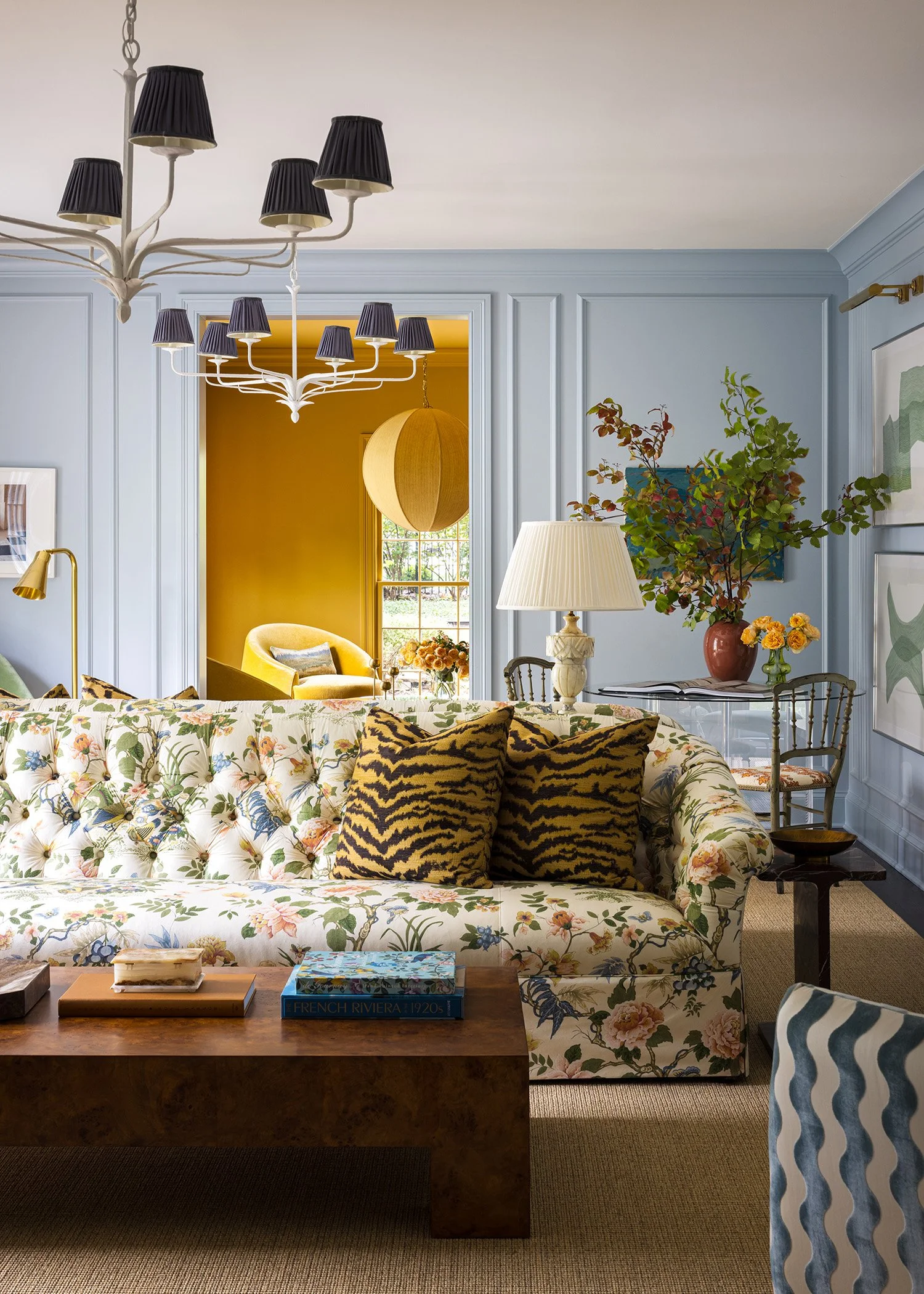 Living room with light blue walls, a floral-patterned sofa with animal print pillows, a wooden coffee table with books, a white chandelier with black lampshades, a large beige lampshade, a potted plant with green and reddish leaves, framed artwork, and a yellow armchair visible through an entryway with warm yellow walls.